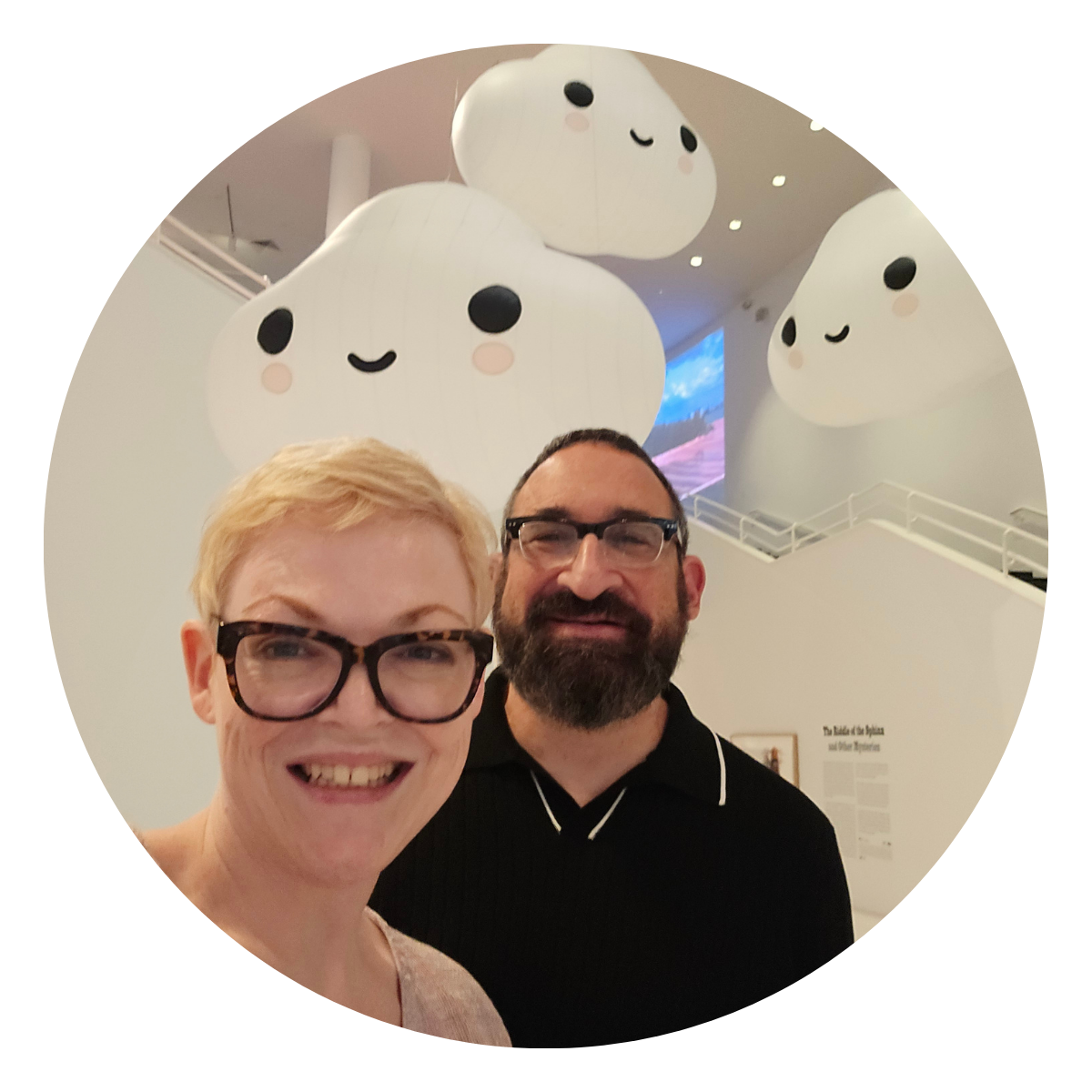 A smiling woman with short blonde hair and glasses, and a man with dark hair, beard, and glasses, taking a selfie in front of three large, cute, smiling cloud sculptures with faces, in a gallery or museum space.
