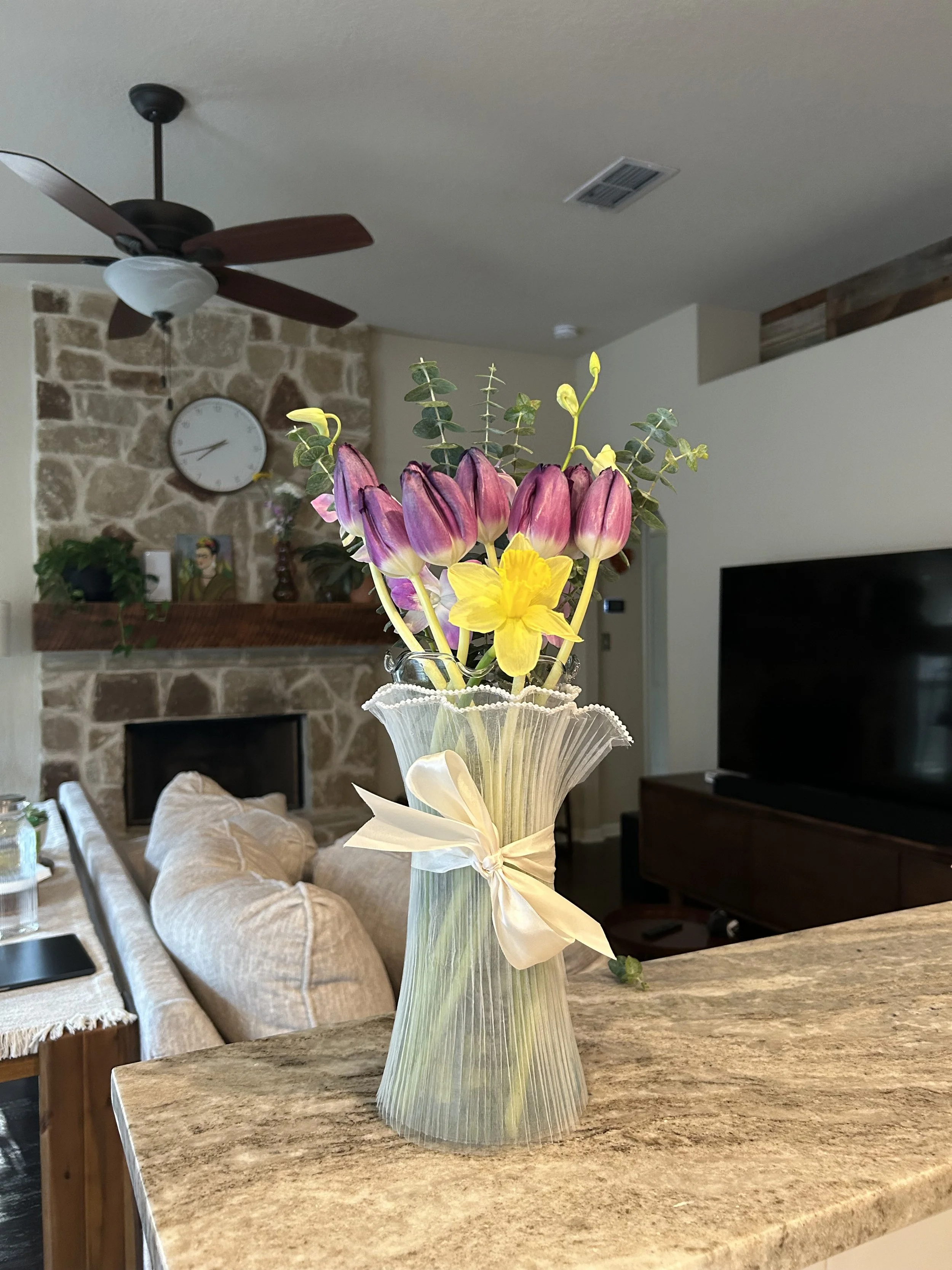 A bouquet of pink, yellow, and purple flowers with green leaves in a clear, pleated glass vase on a kitchen counter.