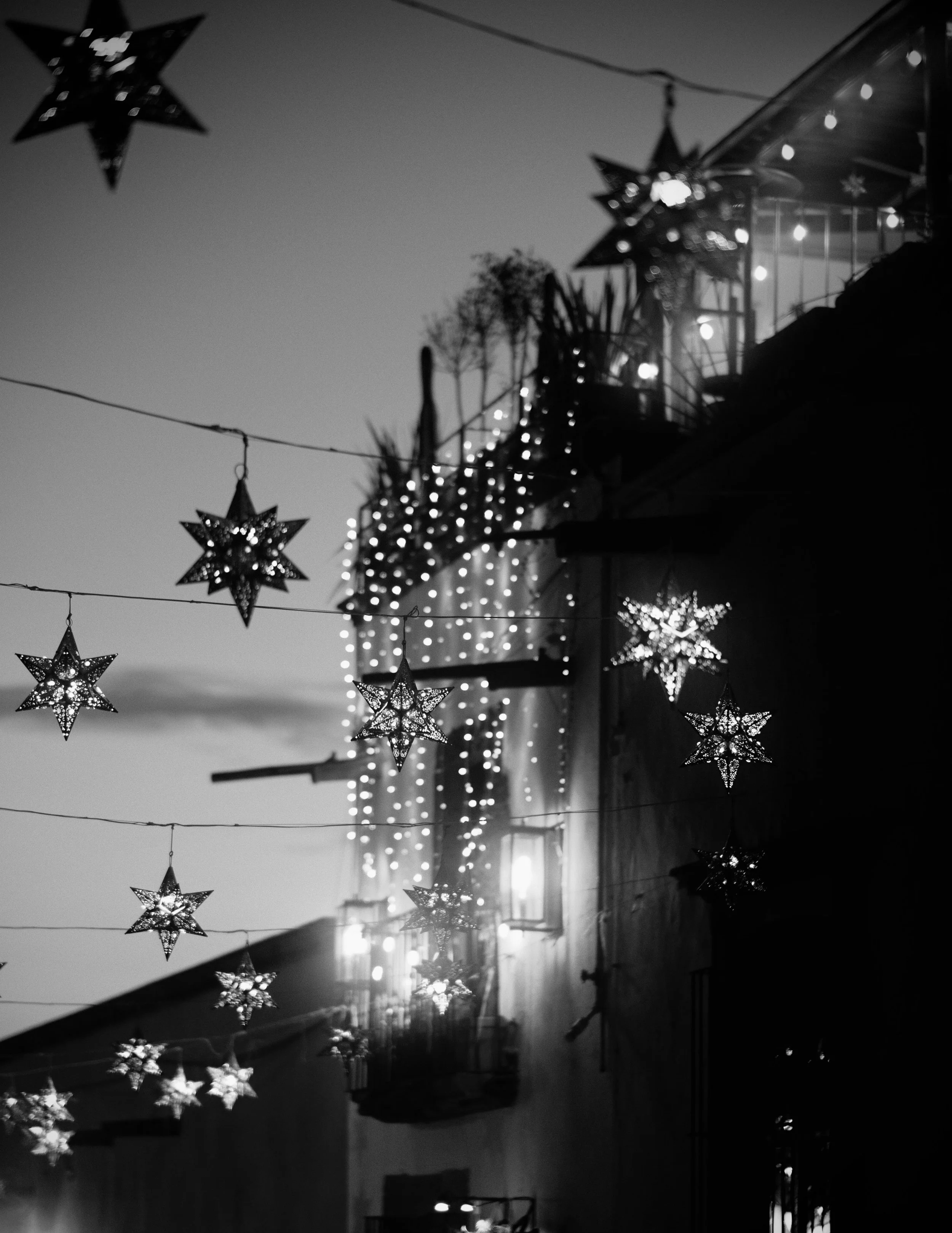 Star-shaped tin lanterns suspend across an evening sky, casting intricate points of light above a softly illuminated facade draped in festival lights. The rich black-and-white tones transform overhead decorations into a floating constellation over th