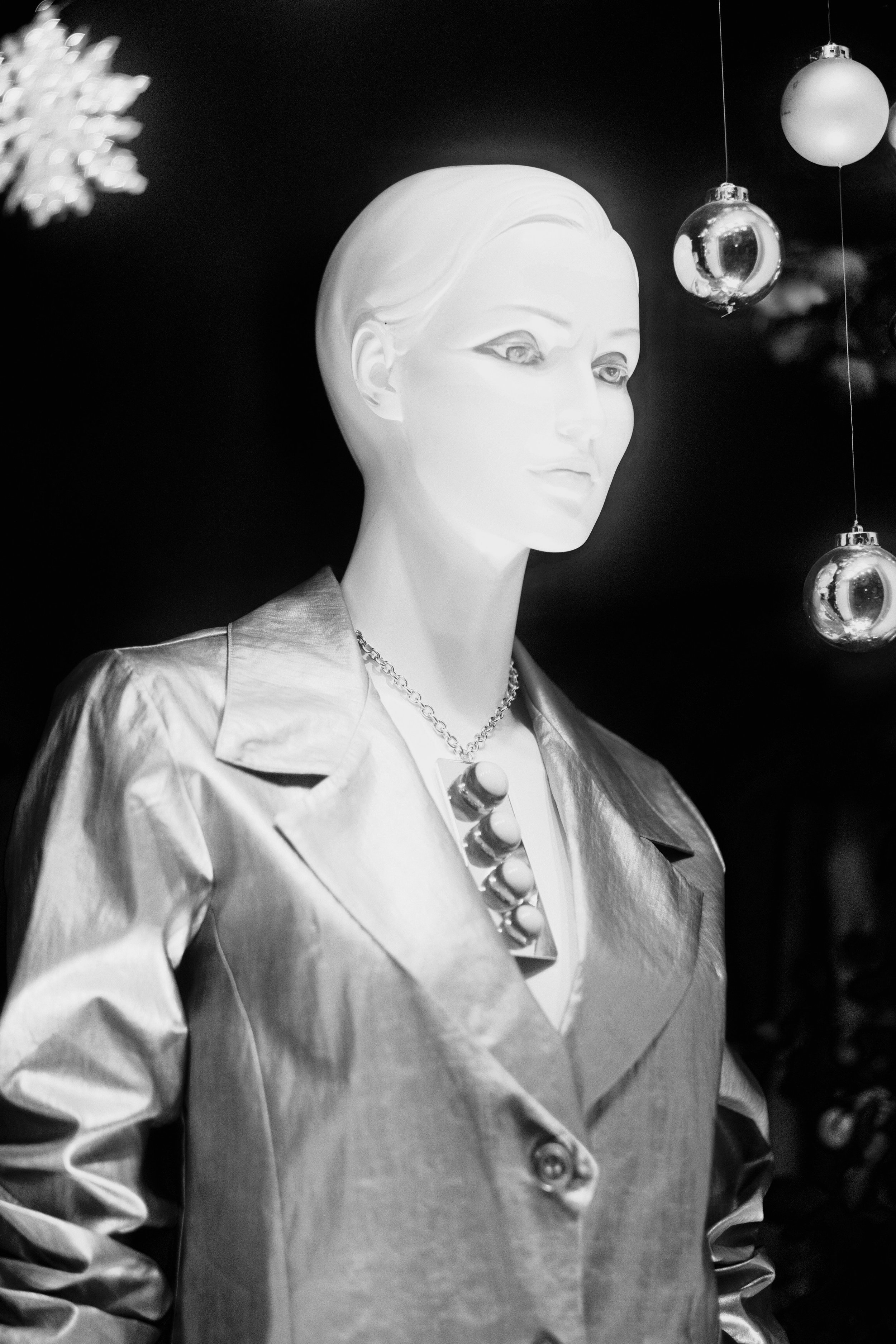 A sleek, sculptural mannequin adorned in a tailored metallic jacket and modern pendant necklace stands suspended against a deep black background, flanked by shimmering holiday ornaments. Sharp directional lighting highlights clean geometric folds and