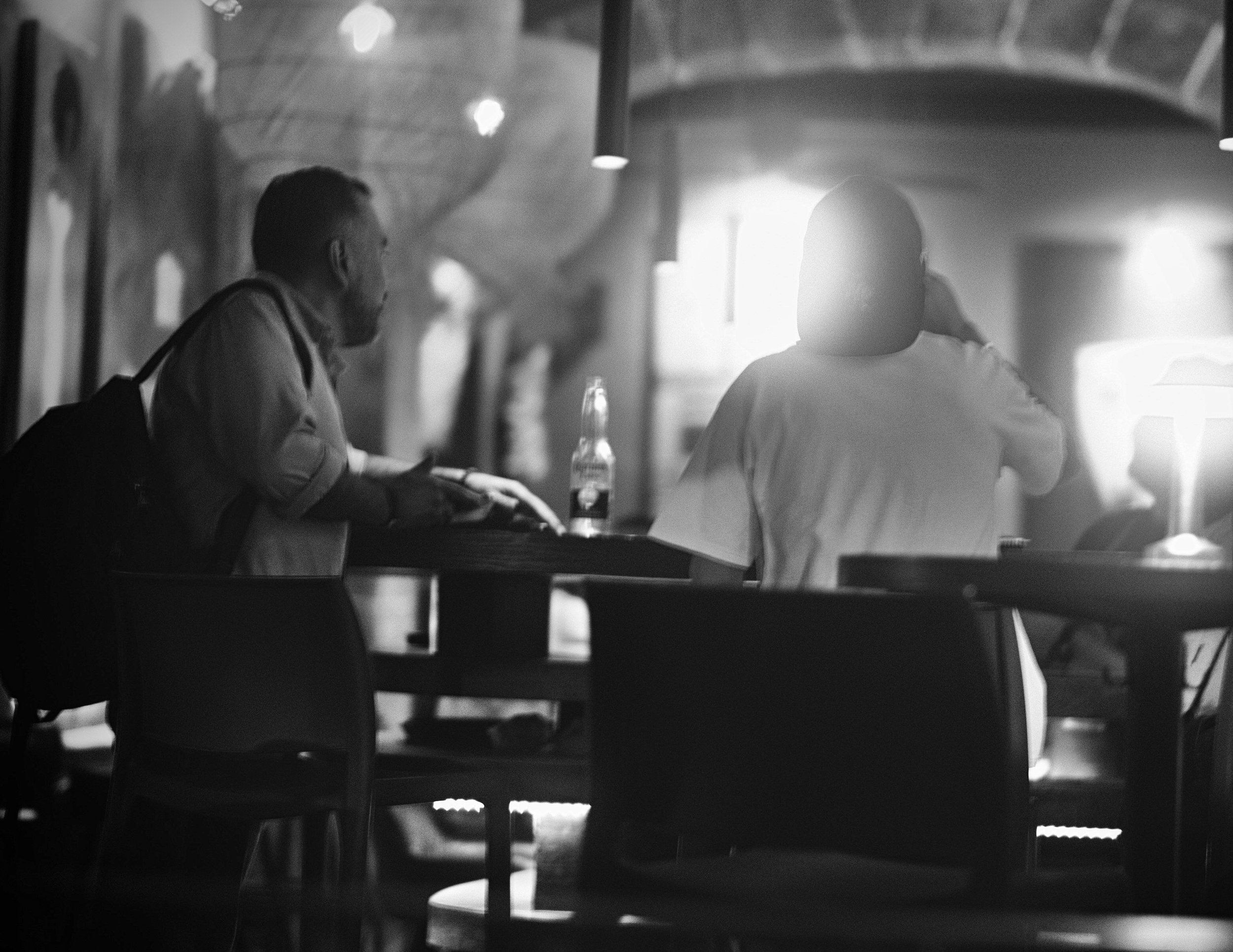 Silhouetted figures linger around a low-lit bar top, bathed in ambient backlighting and soft atmospheric glow. The subtle focus and rich tonal range evoke the quiet, intimate camaraderie of a late-night holiday exchange in San Miguel de Allende.