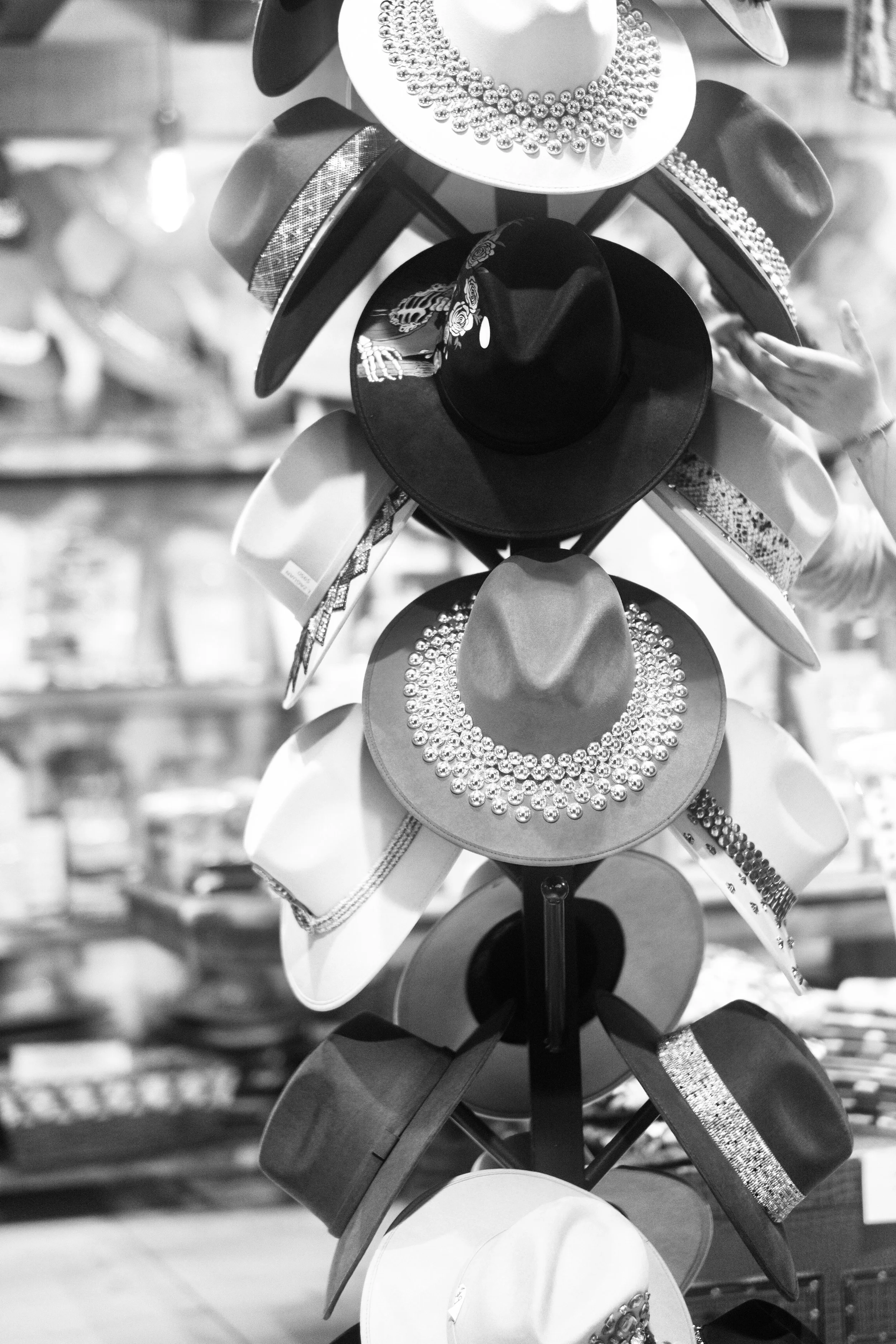 An array of embellished brimmed hats spirals up a display rack, each featuring detailed hatbands, metallic studs, and painted artwork. High-contrast monochrome turns a market display into a rhythmic study of geometry, texture, and local craftsmanship