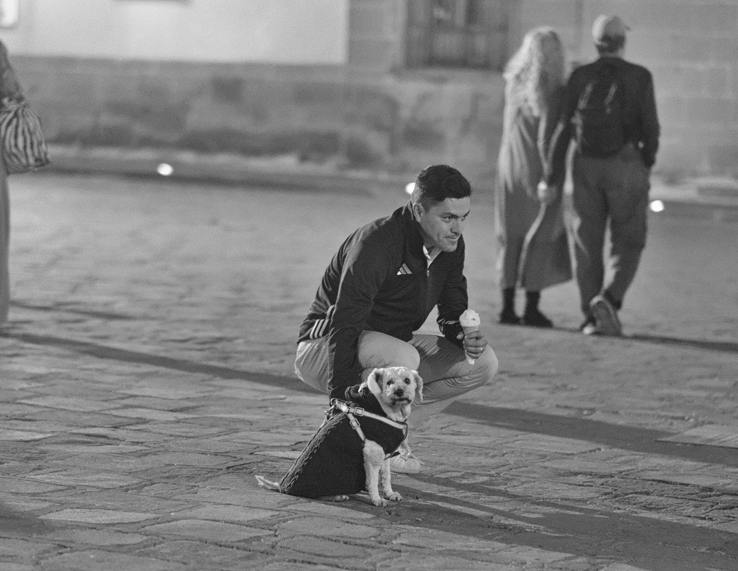 A crouched man enjoying a soft-serve cone rests his hand gently on a small, sweater-clad dog sitting patiently on the stone pavement. Softly blurred figures walking in the background frame a charming, unscripted moment of evening companionship.