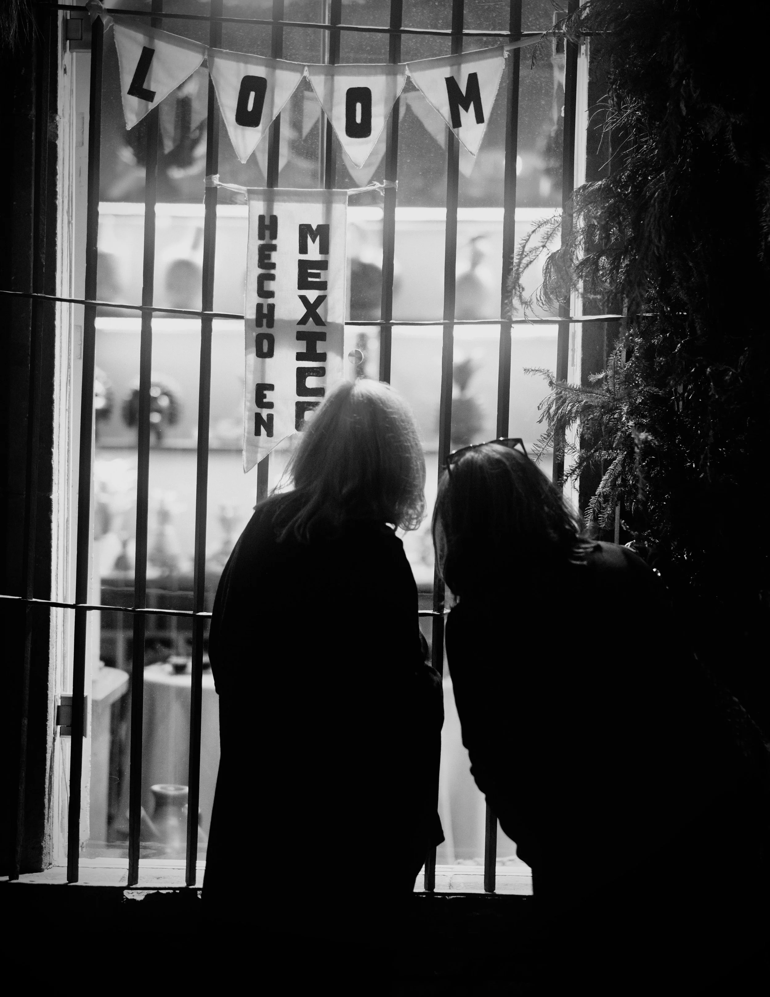Two figures stand silhouetted against a softly illuminated storefront window, peering through iron rejas past hanging banners reading "LOOM" and "HECHO EN MEXICO." The stark monochrome interplay of light and shadow captures a quiet, intimate pause al