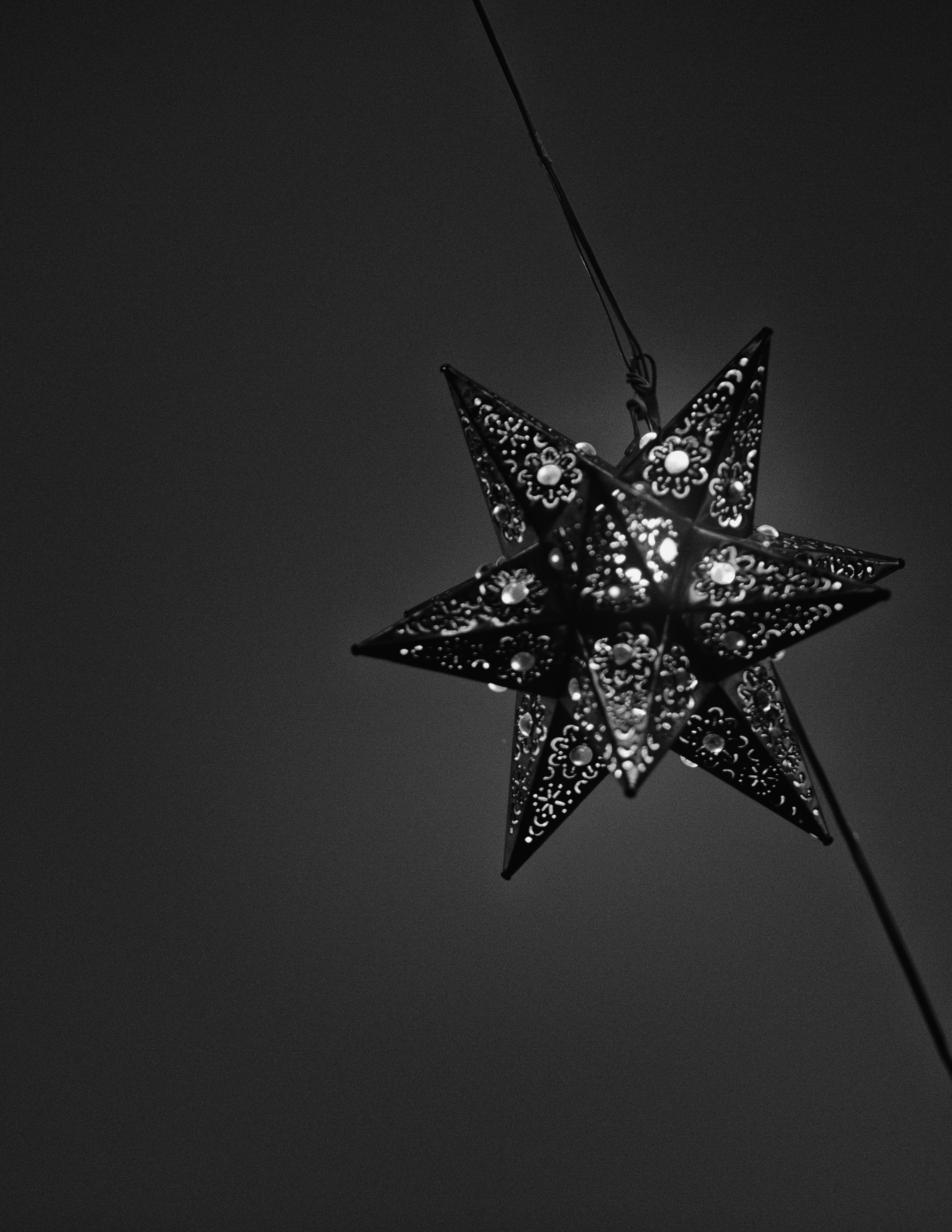 A single handcrafted tin star lantern hangs suspended against a deep, twilight darkness. Delicate perforations and glass embellishments catch subtle ambient light, emphasizing the intricate metalwork and minimal geometric elegance of a classic San Mi