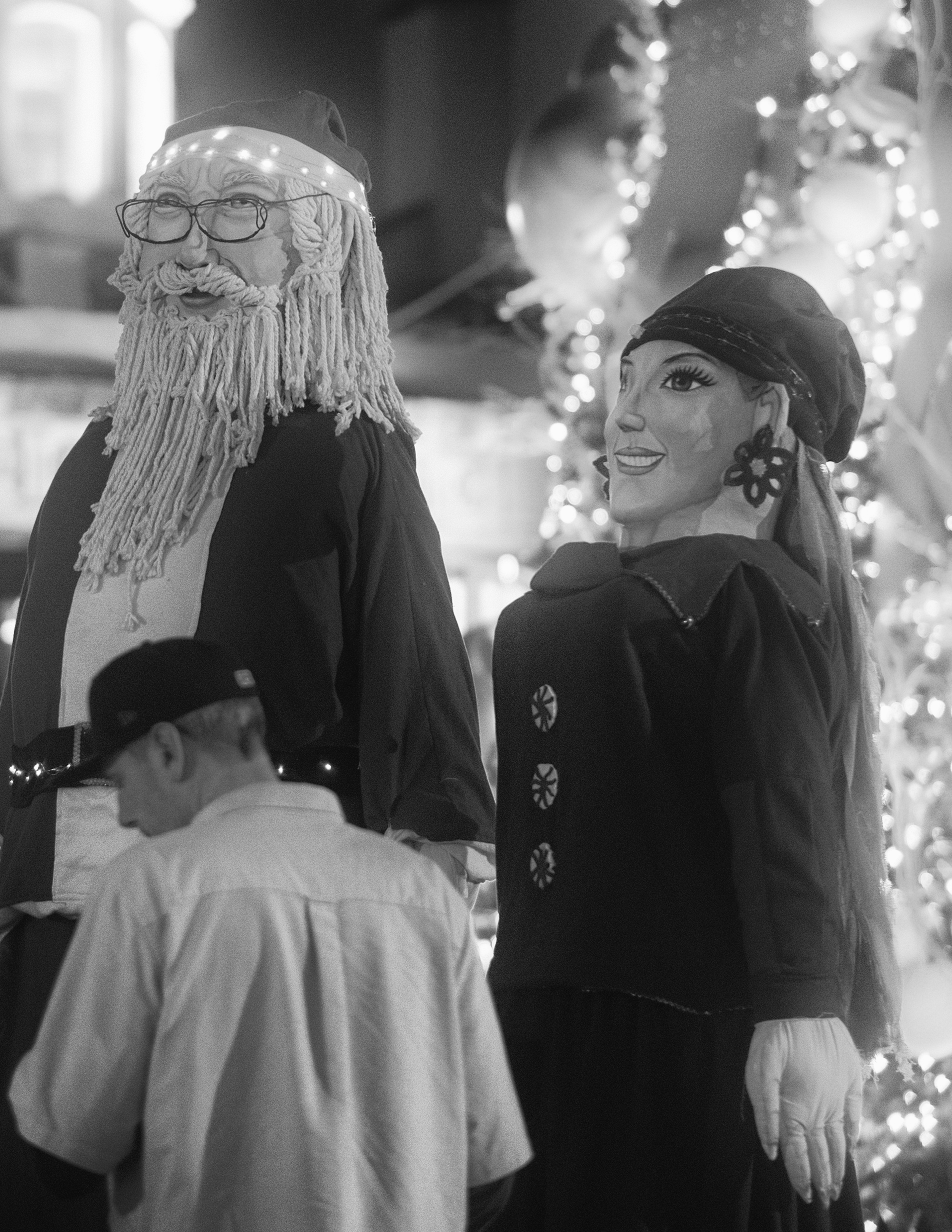 Larger-than-life Christmas Mojigangas—representing Santa Claus and Mrs. Claus—tower over evening passersby amidst a wash of festive bokeh lights. The shot juxtaposes traditional Mexican papier-mâché artistry with holiday iconography, capturing the wh