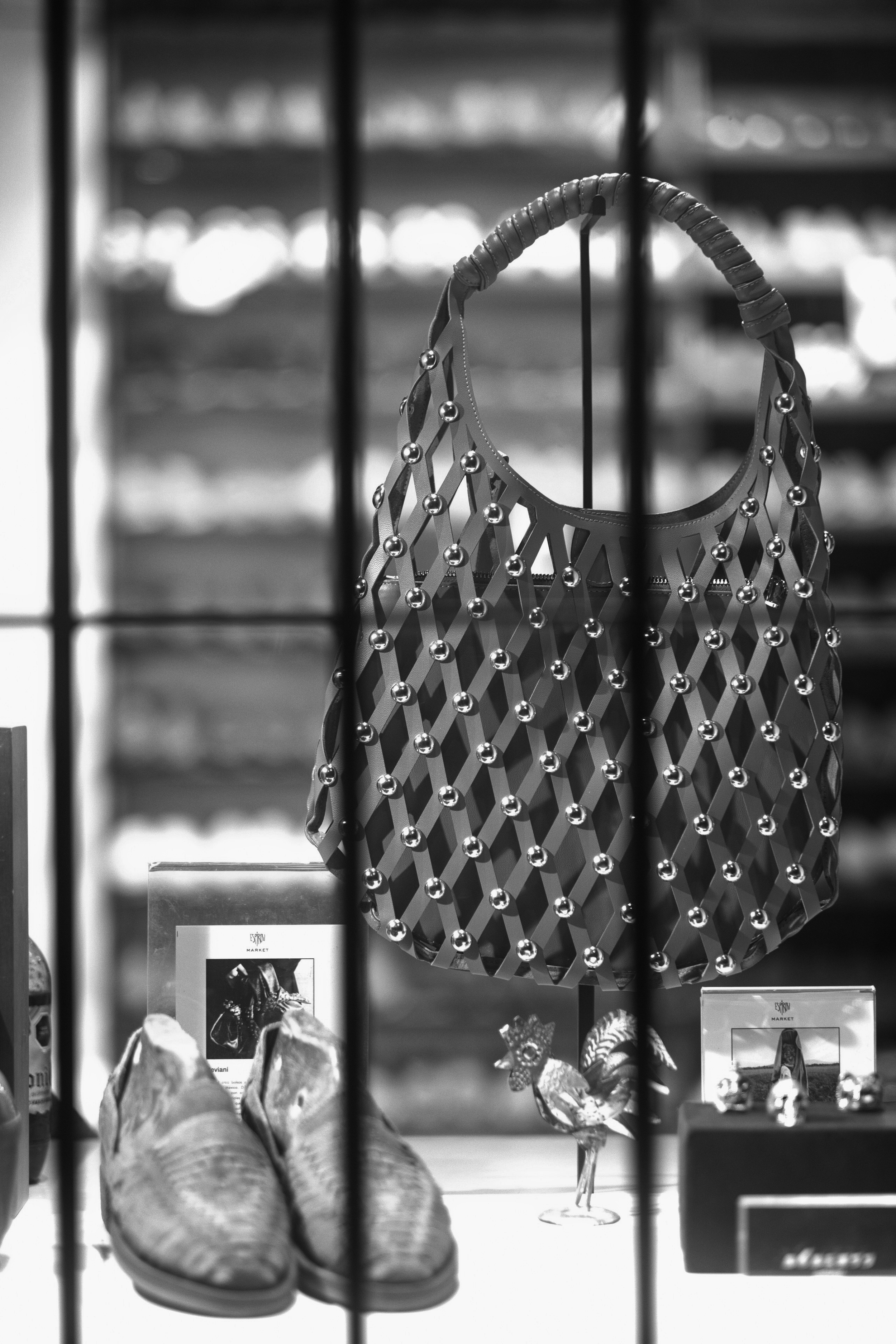 An intricately woven leather handbag with metallic studs takes center stage in a lighted boutique display, anchored by handcrafted shoes and small artisanal details below. Framed behind vertical iron bars, the image plays on overlapping geometric pat