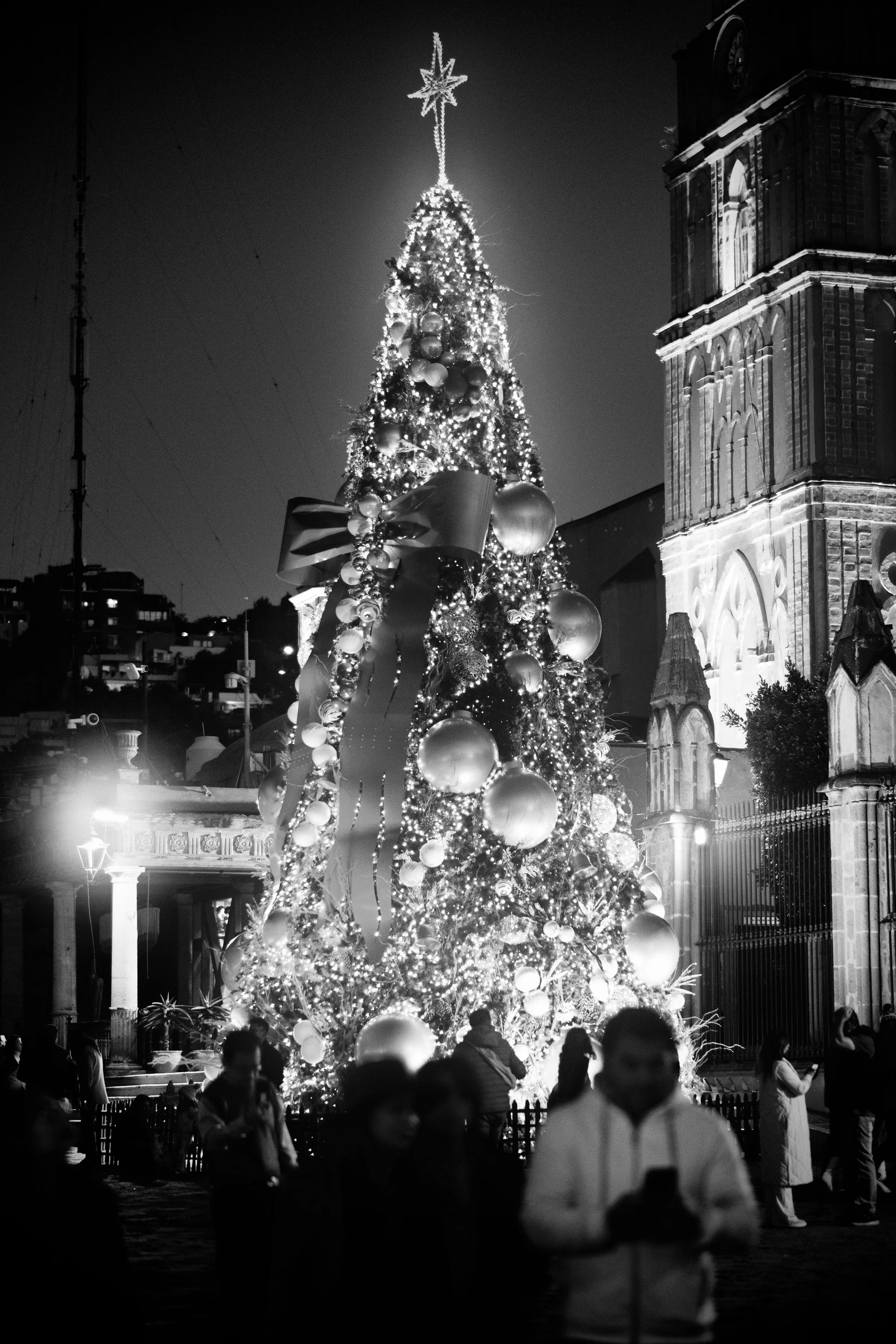 A towering, brilliantly lit Christmas tree anchored by a glowing star topper dominates the central plaza, set against the silhouette of San Miguel's iconic parish architecture. Figures drift through the foreground in soft motion, capturing the lumino