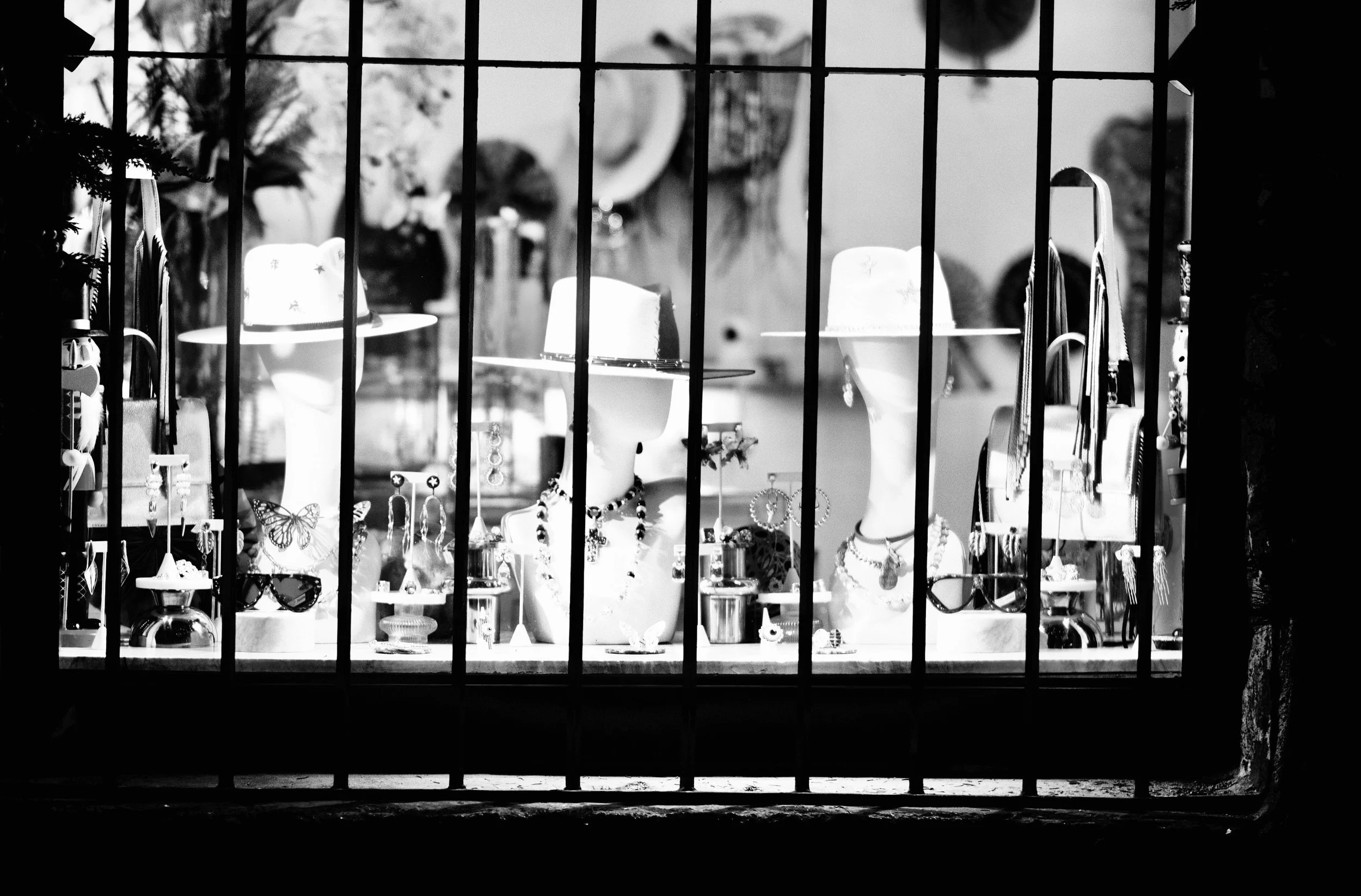 Three hat-wearing mannequins and delicate jewelry displays are illuminated within a storefront window, framed strictly by vertical iron bars. High-contrast monochrome creates a striking graphic rhythm, turning an after-hours shop window into a quiet 