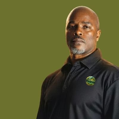 A man with dark skin and a shaved head, wearing a black collared shirt with a green button or badge, standing against an olive green background.