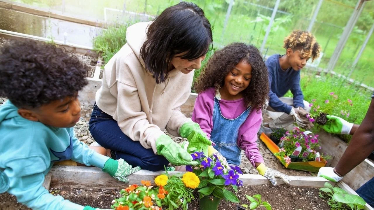 A woman and children planting flowers in a greenhouse garden. Links to farm based summer camp activities.