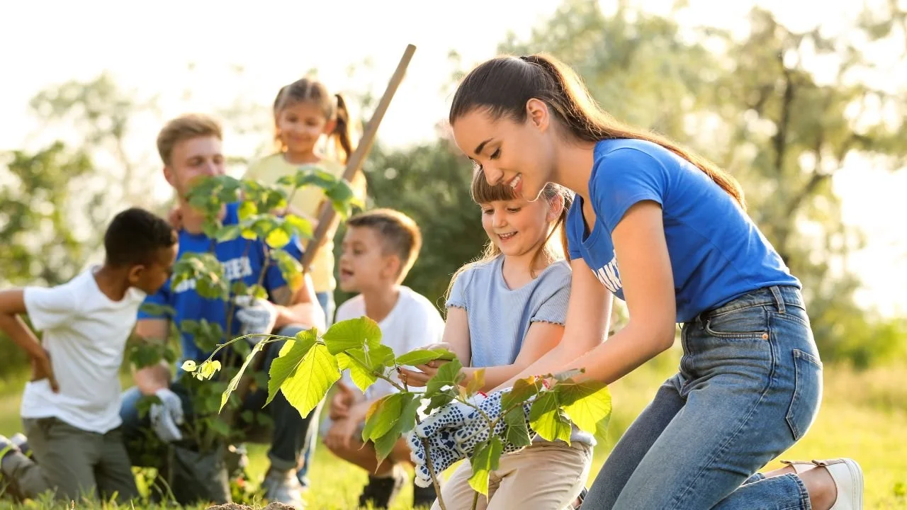 Group of children and a young woman planting a tree outdoors on a sunny day. Links to agri-learning summer camp experiences near Augusta.