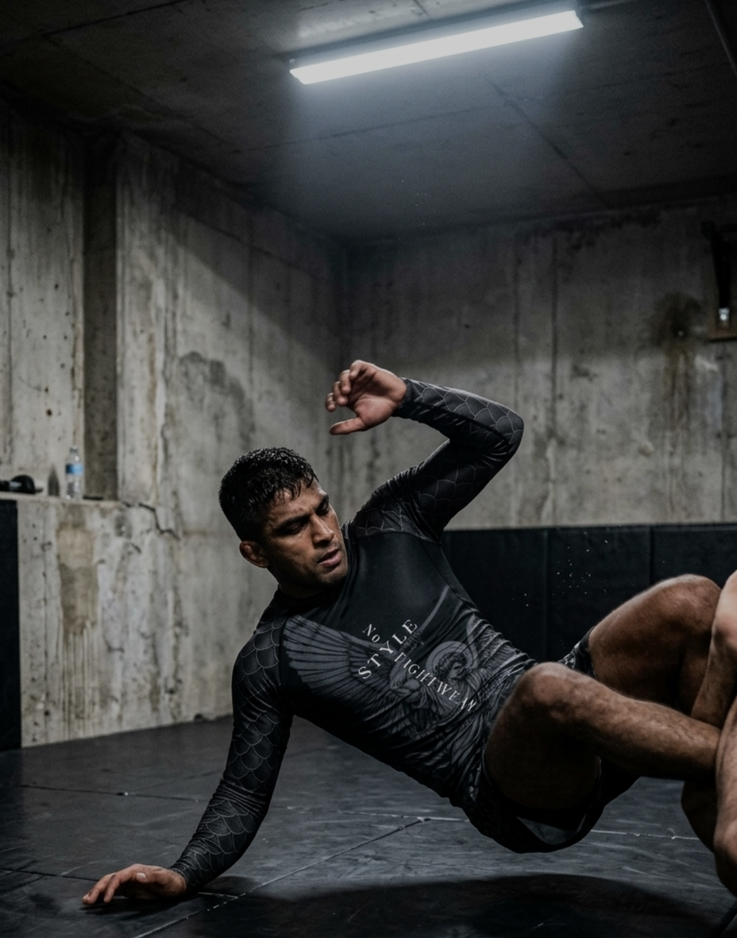 A male athlete practicing Brazilian Jiu-Jitsu on a black mat in an indoor training room with concrete walls and overhead fluorescent lighting.