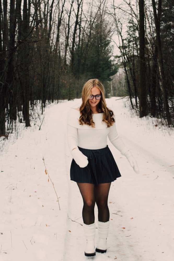 A young woman with glasses smiling, dressed in a white sweater, black skirt, black tights, white winter boots, and white gloves, standing on a snowy forest path.