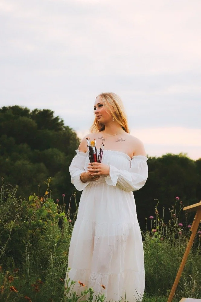 A woman with blonde hair wearing a white off-shoulder dress, holding paintbrushes, standing outdoors in a field with green plants and flowers, under a cloudy sky.