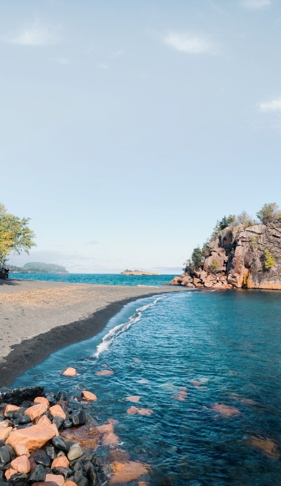 A natural landscape showing a river or creek flowing into a larger body of water at a rocky beach with small waves, a rocky cliff on the right, and trees on the left under a partly cloudy sky.