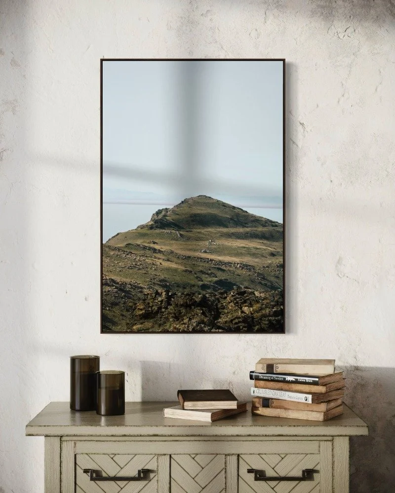 A framed landscape photo of a rugged hillside hangs on a white wall above a vintage dresser. The dresser holds several books, some stacked and some laying flat, with two dark-colored candles on the left.
