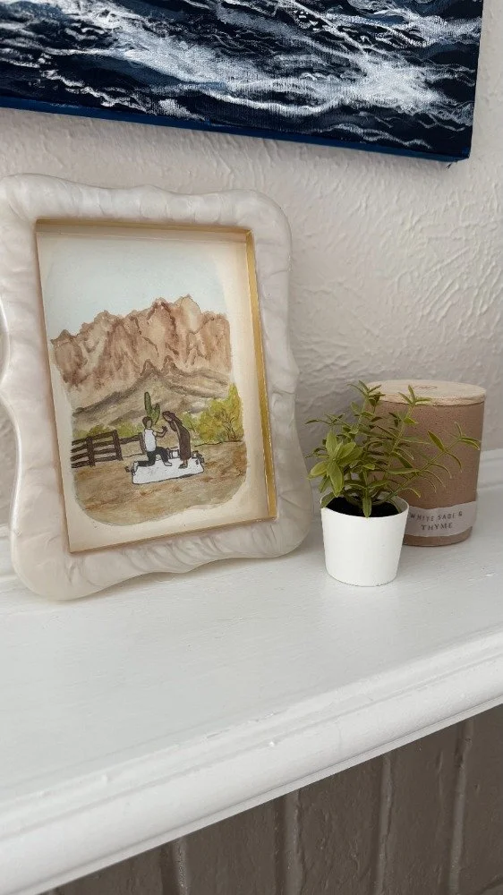 Watercolor painting of two figures with a cactus and mountain background, framed in a textured white frame, placed on a white shelf next to a small potted plant and a round container.