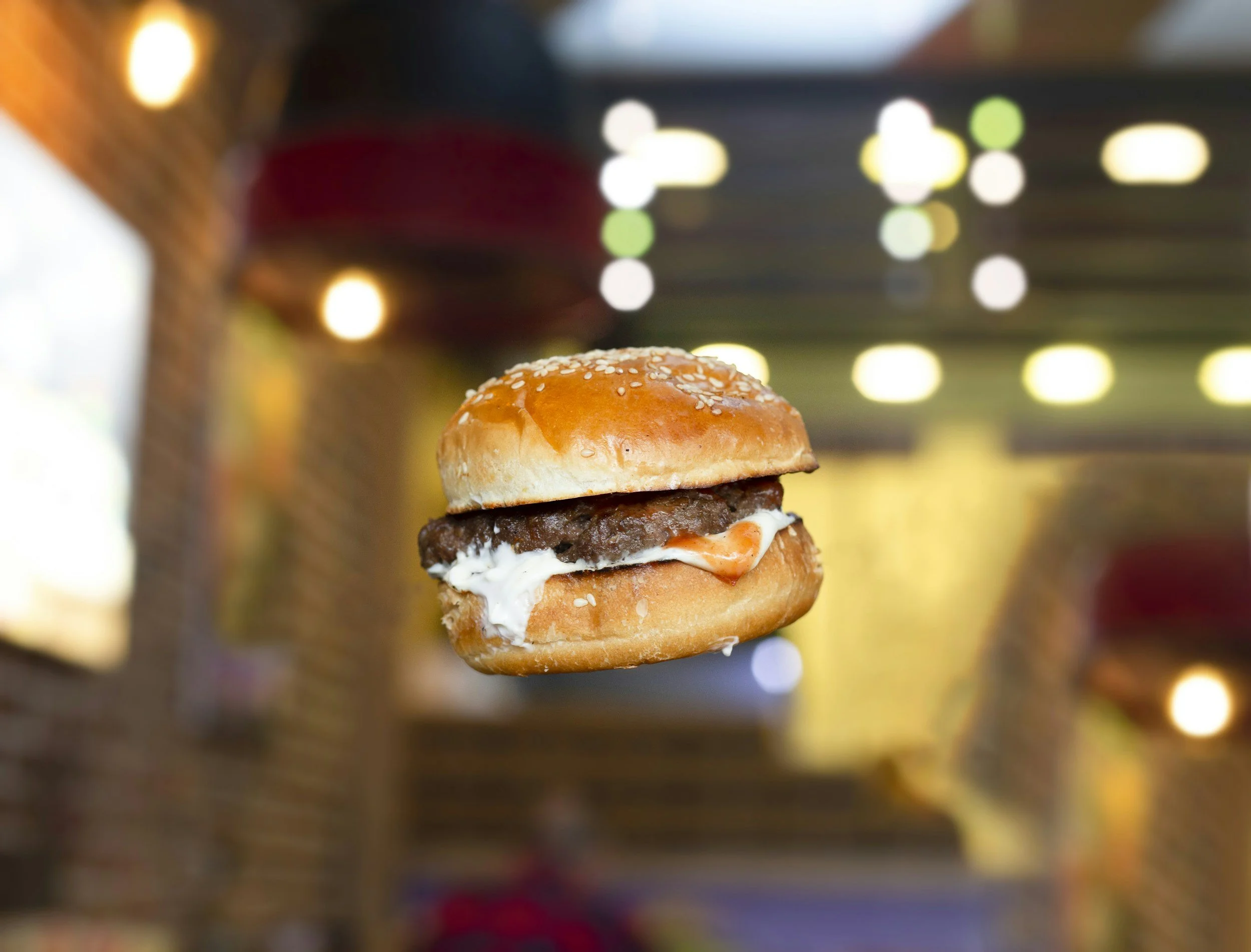 Close-up of a hamburger with sesame seed bun, beef patty, and condiments, floating in the air with blurry colorful lights in the background.