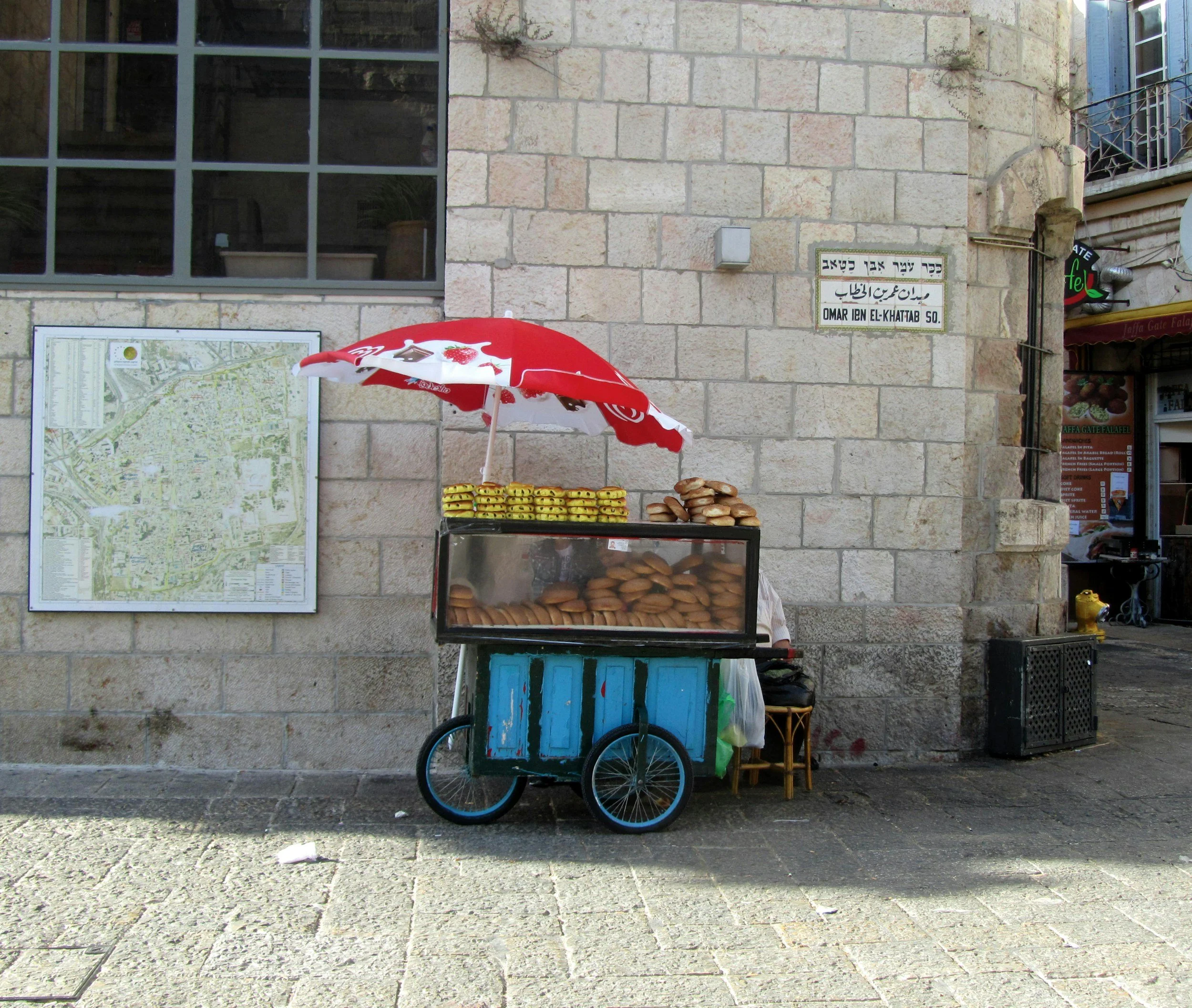 A street vendor cart selling baked goods, including cookies and bread, is positioned in front of a stone building with windows and street signs in an urban area.