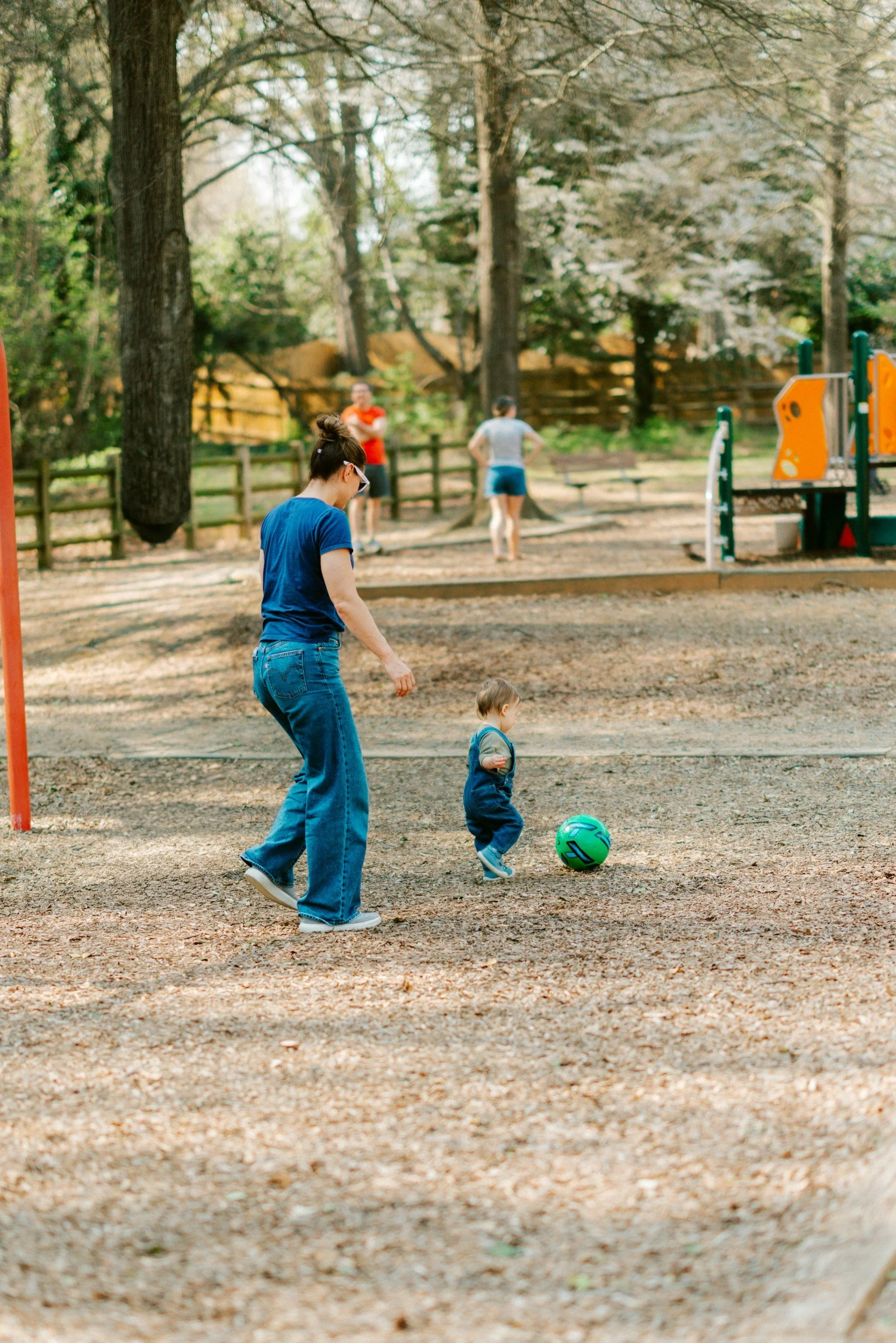 A woman and a young child are playing soccer outdoors on a dirt field, surrounded by trees and a playground in the background.