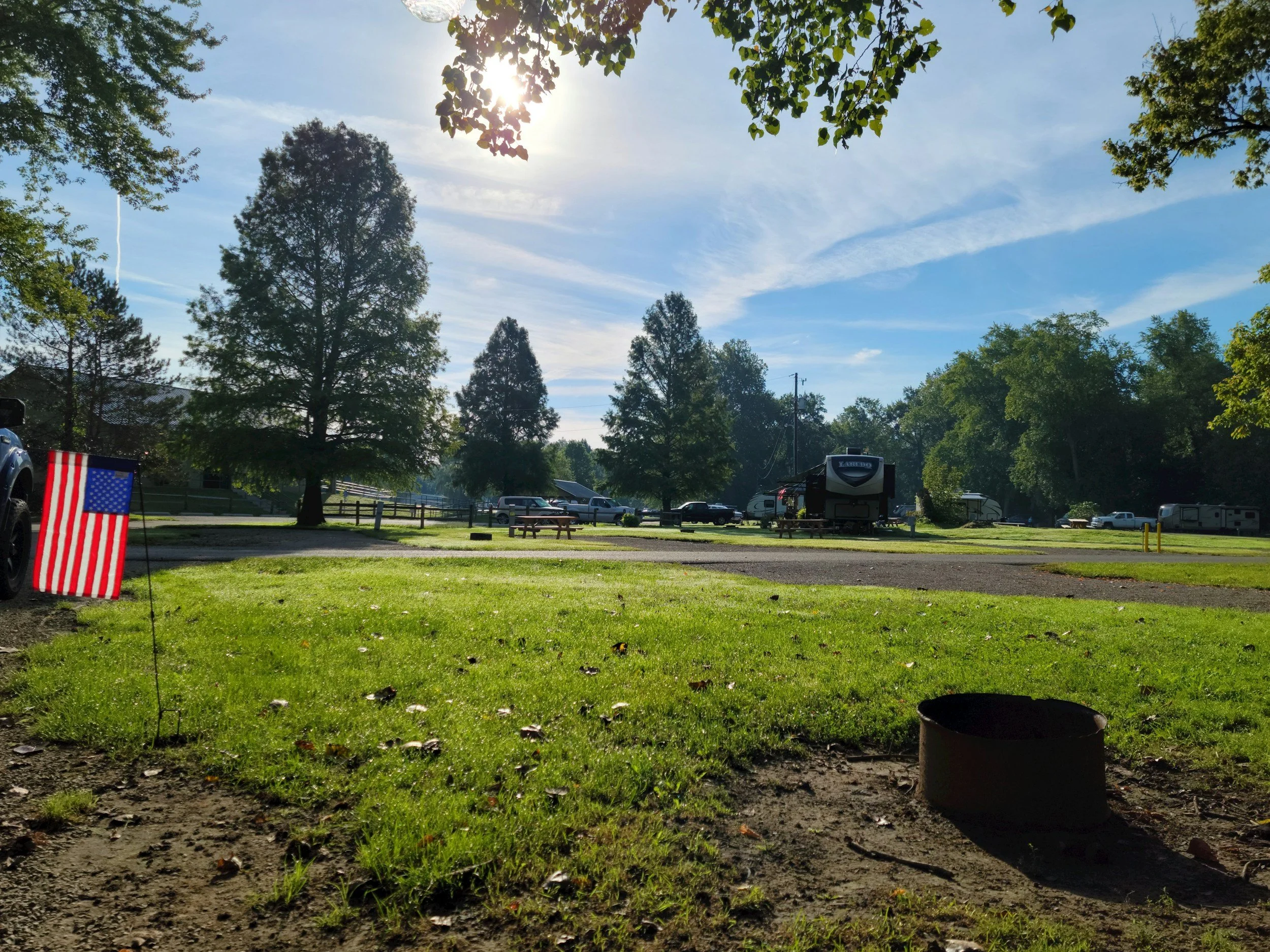 Sun shining through trees over a park with cars, RV, picnic tables, and an American flag.