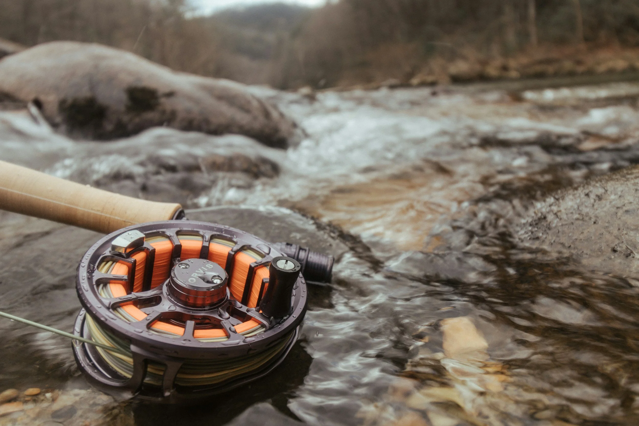 A fishing reel with line on a rock next to a flowing stream in a natural outdoor setting.