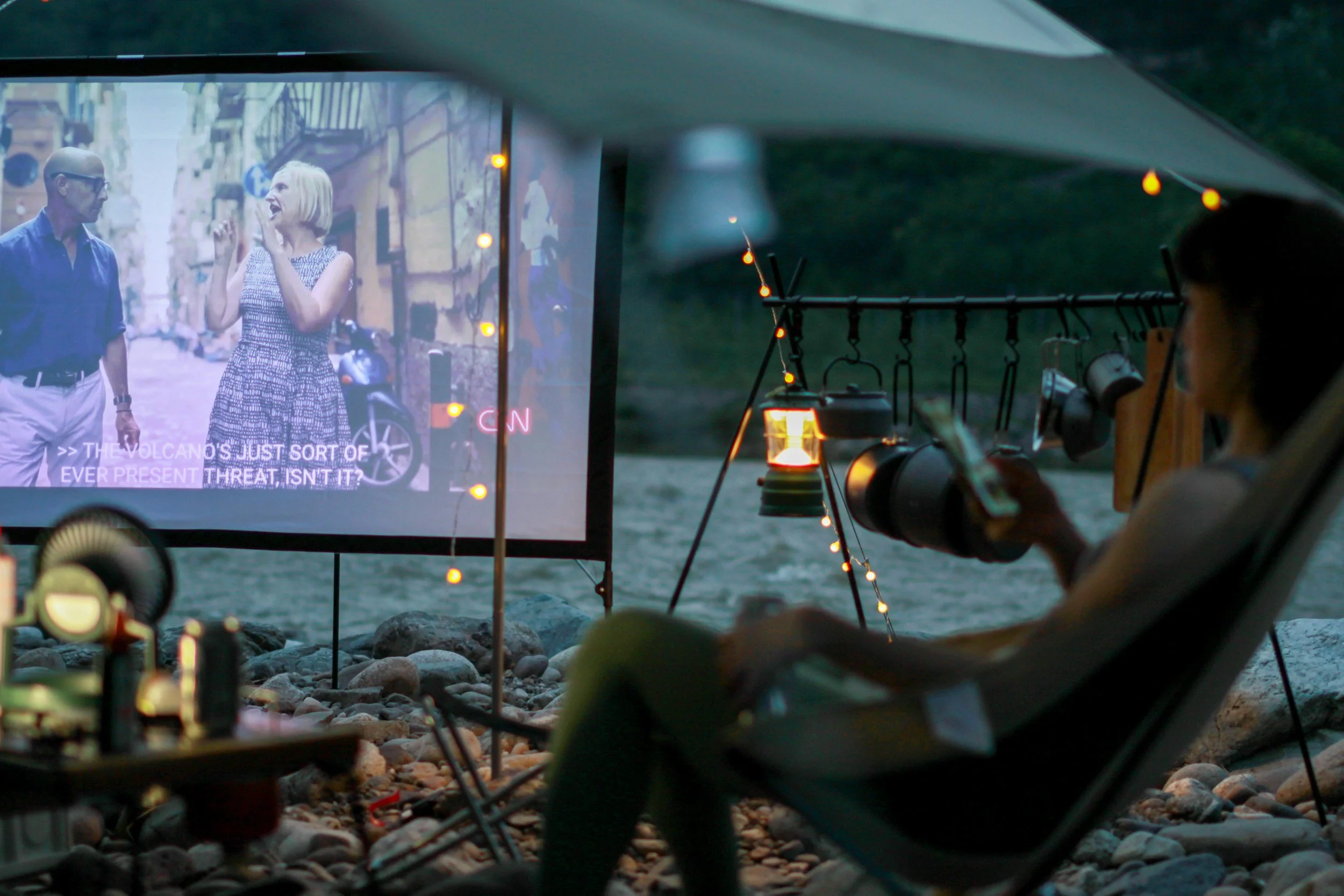View from inside a tent showing a person with a flashlight watching a projected image of a man and woman on a screen outside. The projected scene appears to be part of a presentation or movie with motion graphics and subtitles.