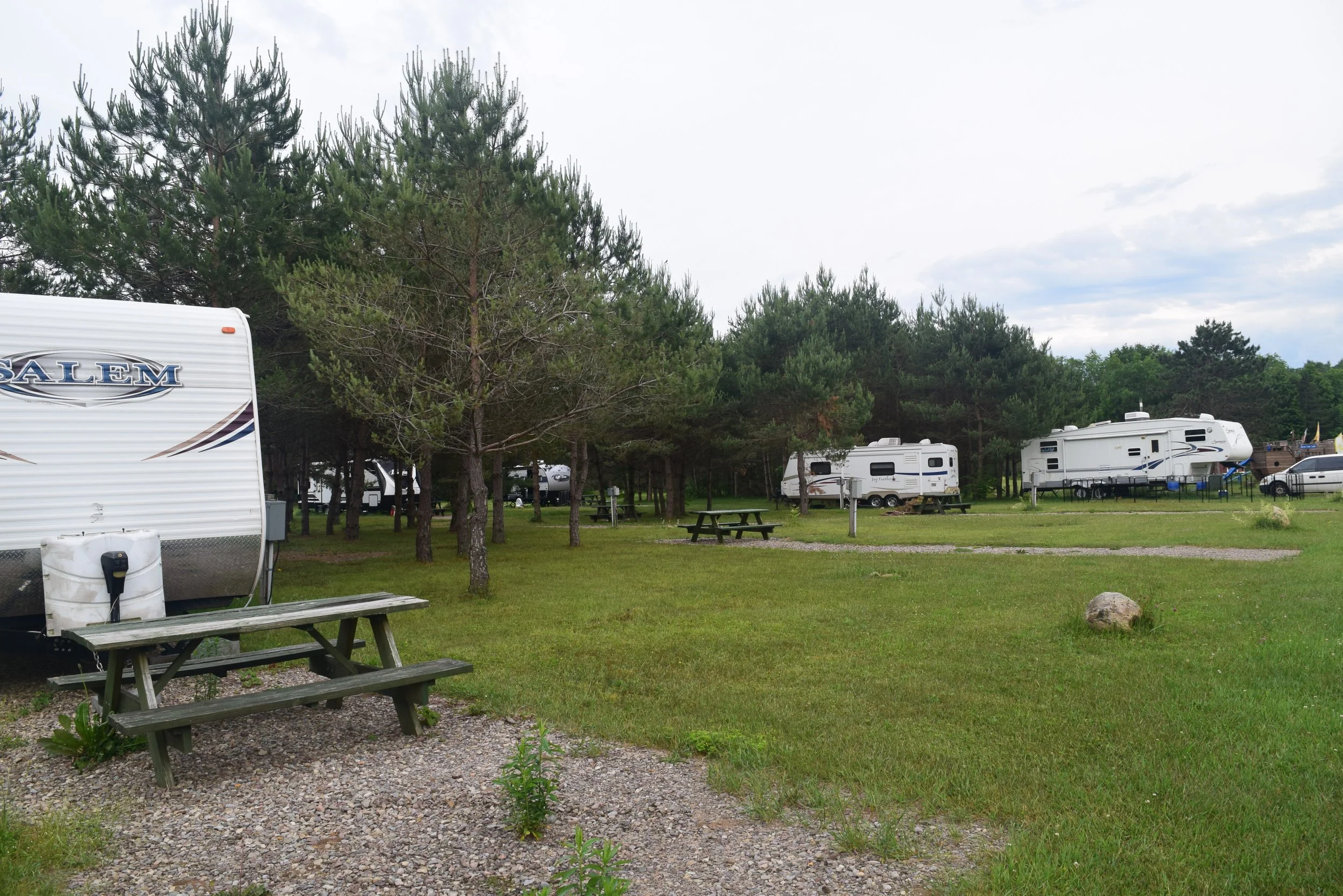Sun shining through trees over a park with cars, RV, picnic tables, and an American flag.