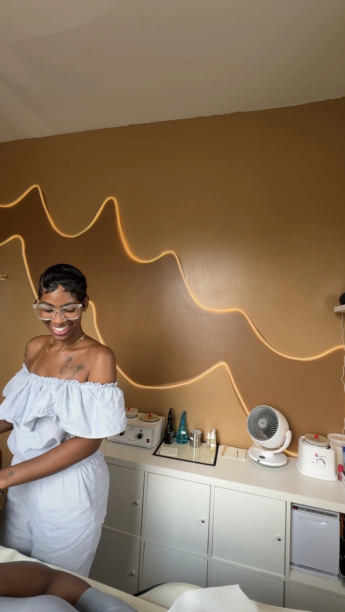 A woman smiling and wearing glasses and a white off-the-shoulder dress standing in a room with a dark brown wall. The wall has a gold neon light in the shape of a mountain range. There is a white cabinet below with various beauty and skincare products, a fan, and an electric towel warmer.
