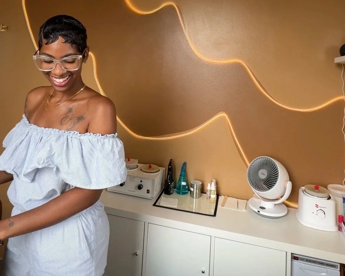 A smiling black woman with short curly hair, glasses, and a tattoo on her chest, wearing a light blue and white off-the-shoulder dress, standing inside a Harlem Brazilian sugaring studio with a decorative gold wavy wall.