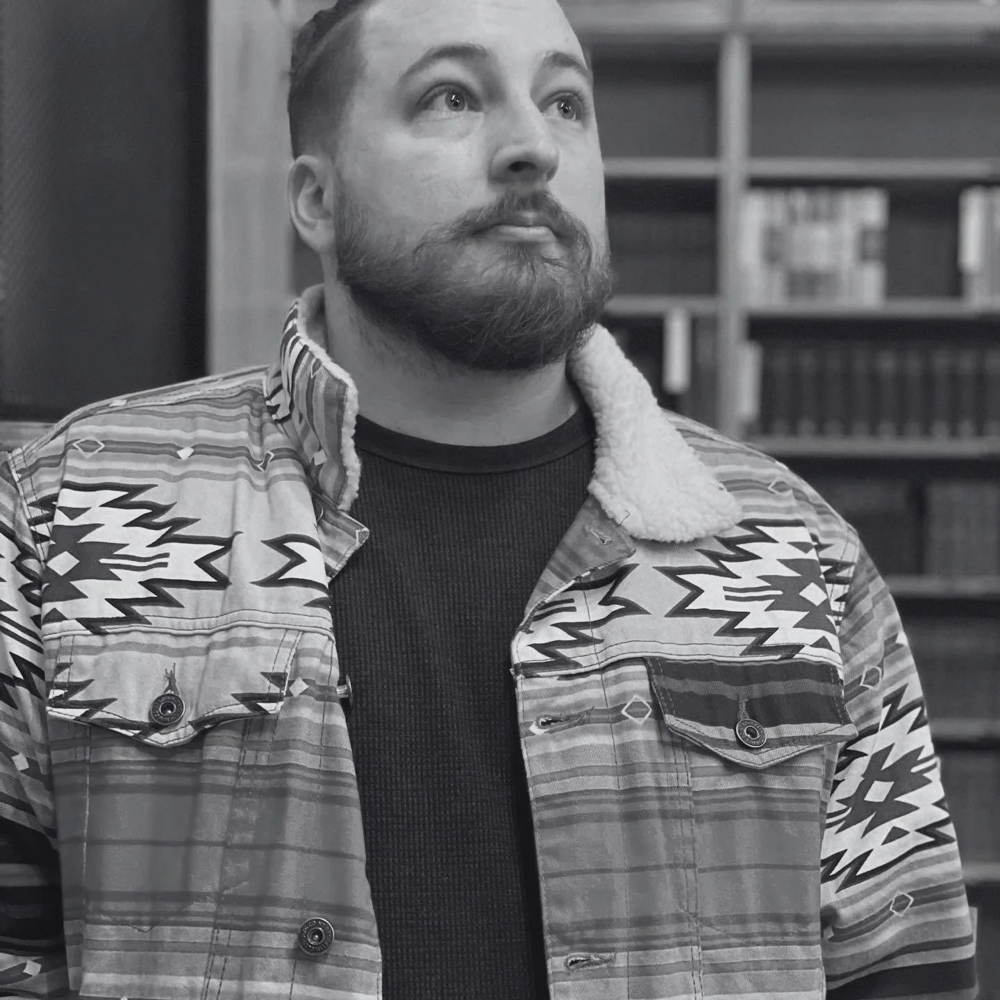 A man with a beard and short hair, wearing a patterned shirt with a shearling collar over a dark shirt, standing indoors with a bookshelf in the background.