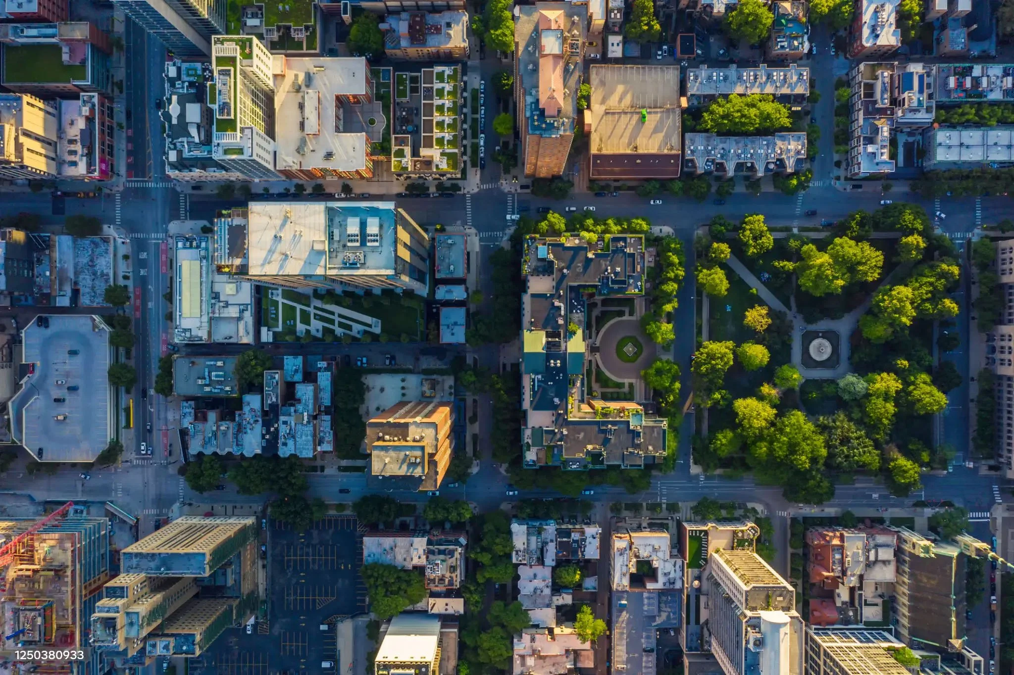 An aerial view of a city grid featuring tall buildings, streets with cars, and plenty of green trees and parks, taken during daylight.