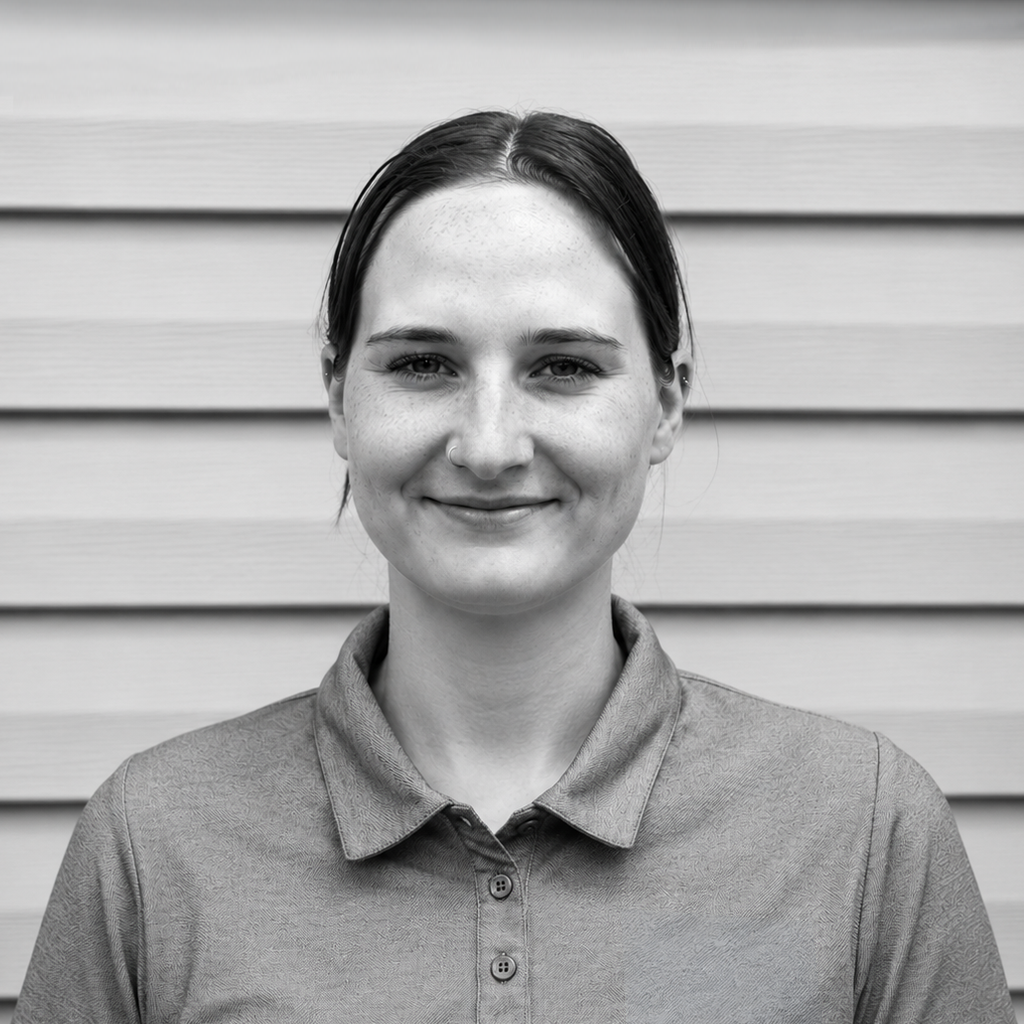 Black and white photo of a young woman with straight hair tucked behind her ears, wearing a polo shirt, standing in front of a wooden wall, smiling gently.