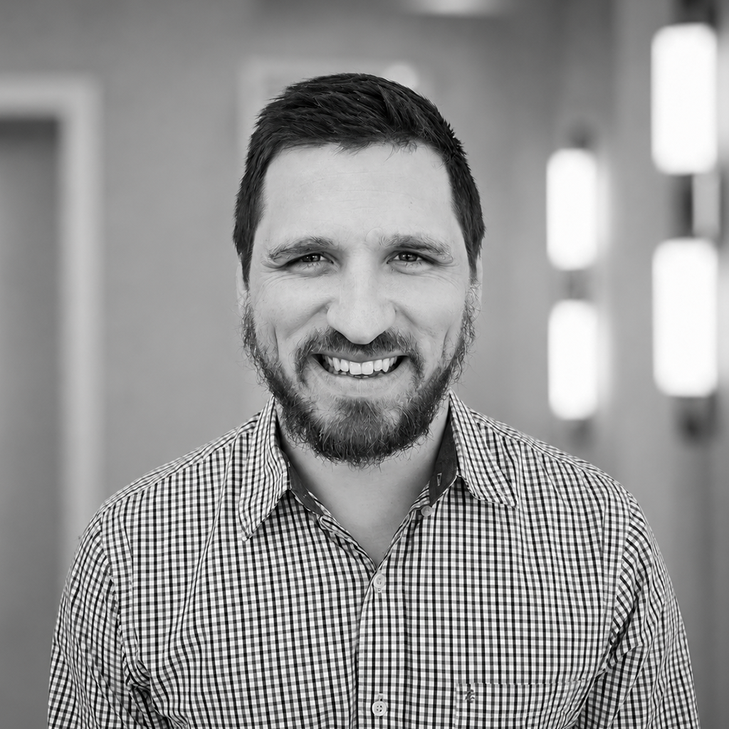 A smiling man with a beard and short hair, wearing a checkered shirt, standing indoors with blurred background of windows or lights.