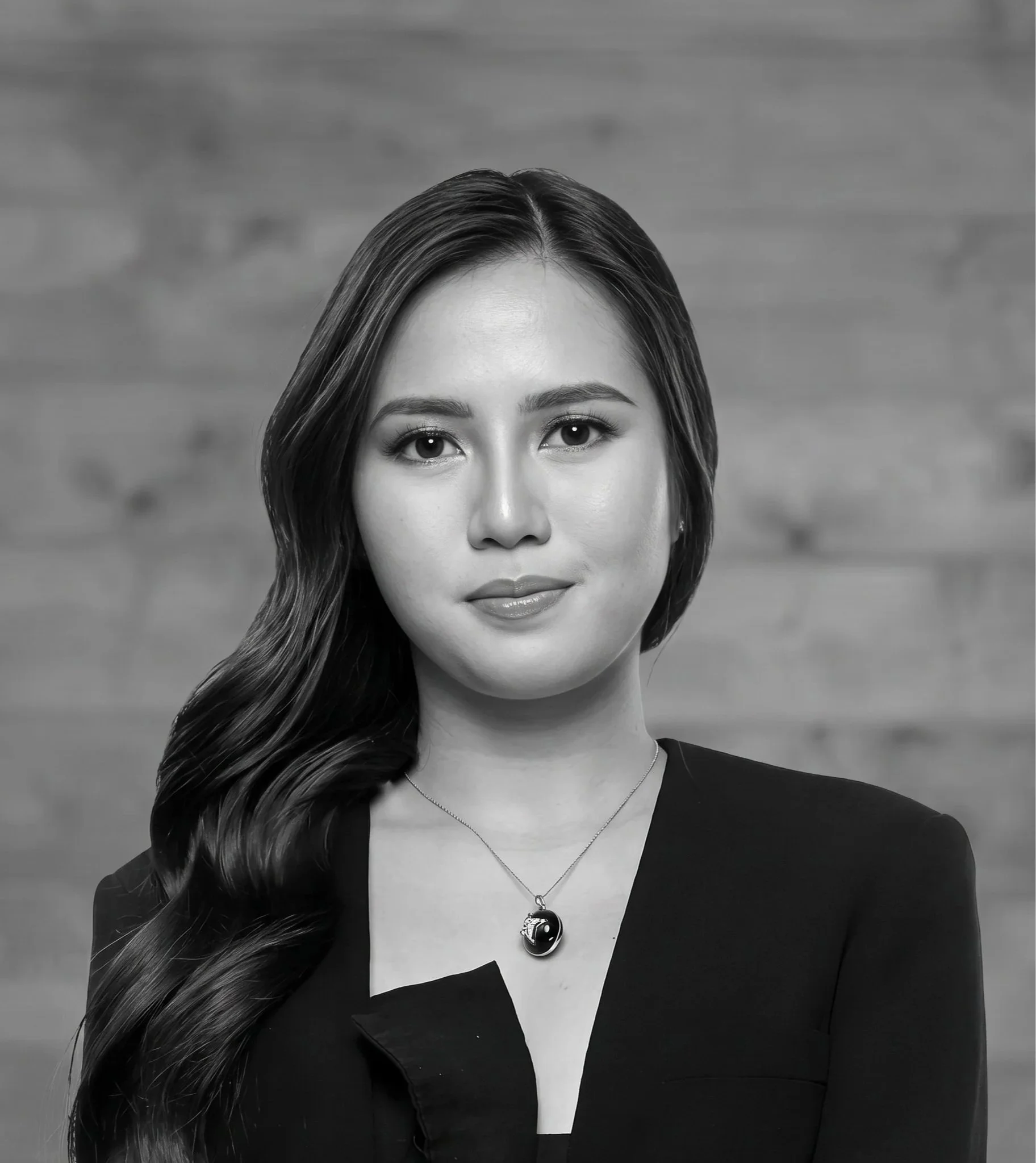 Close-up black and white portrait of a woman with long wavy hair, wearing a necklace with a yin-yang pendant, and a blazer with an open white shirt underneath, standing in front of a blurred brick wall background.