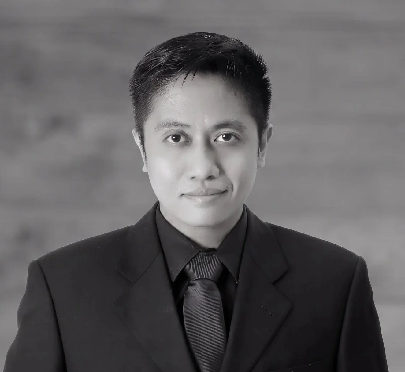 A black-and-white portrait of a young man in a suit and tie, looking directly at the camera with a neutral expression.