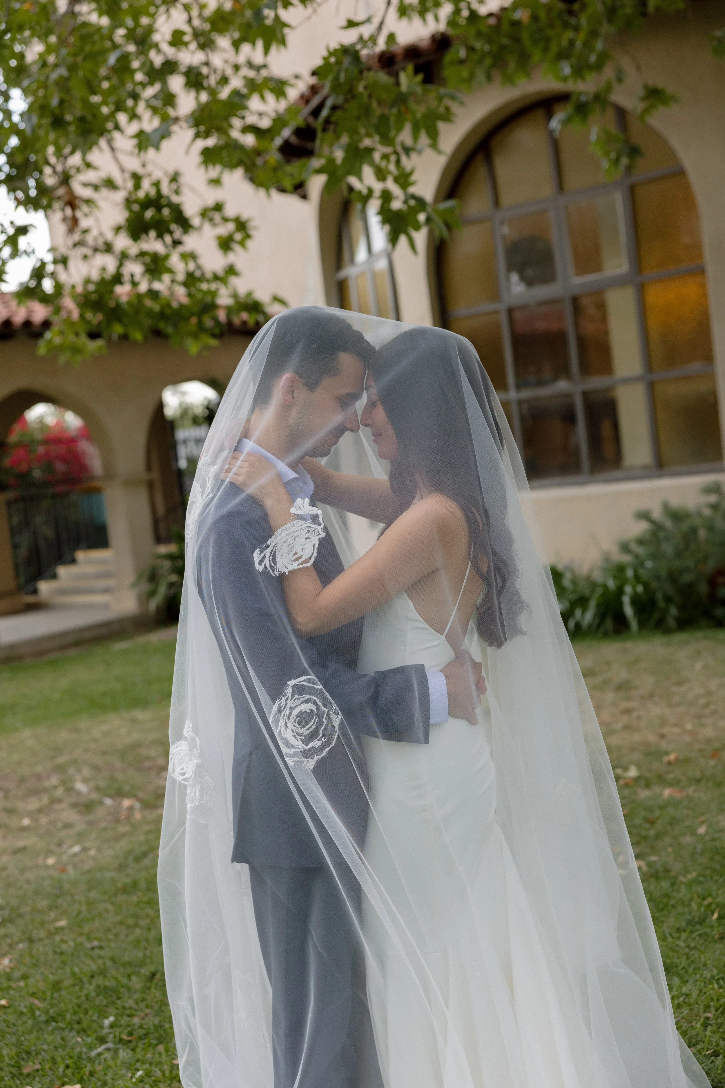 A bride and groom sharing an intimate moment under a wedding veil outdoors near a building with large arched windows, surrounded by greenery.