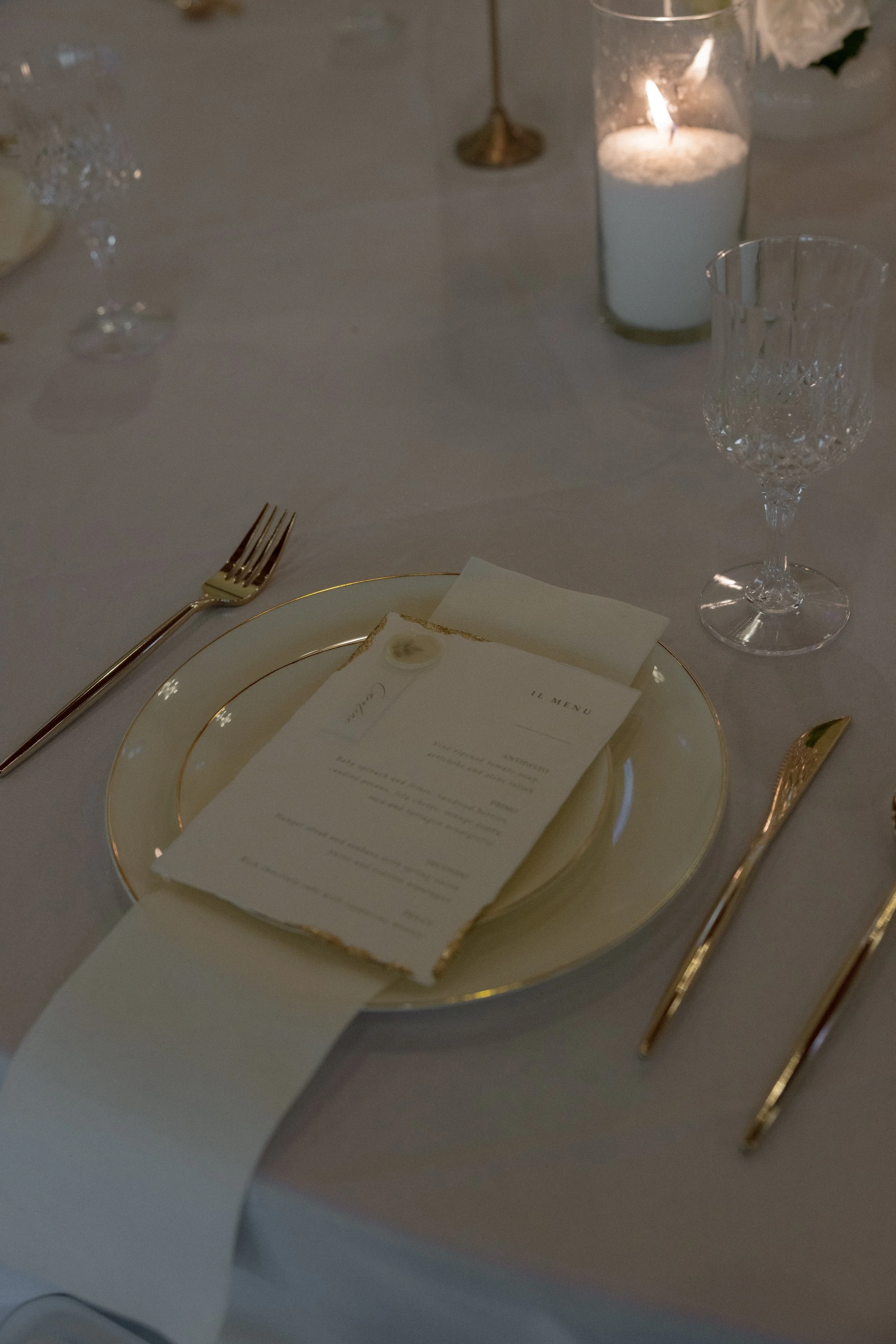 A formal place setting with a gold-rimmed white plate, gold cutlery, and a menu card on top. In the background, there are wine glasses, a paper napkin, a candle in a glass holder, and a tablecloth.
