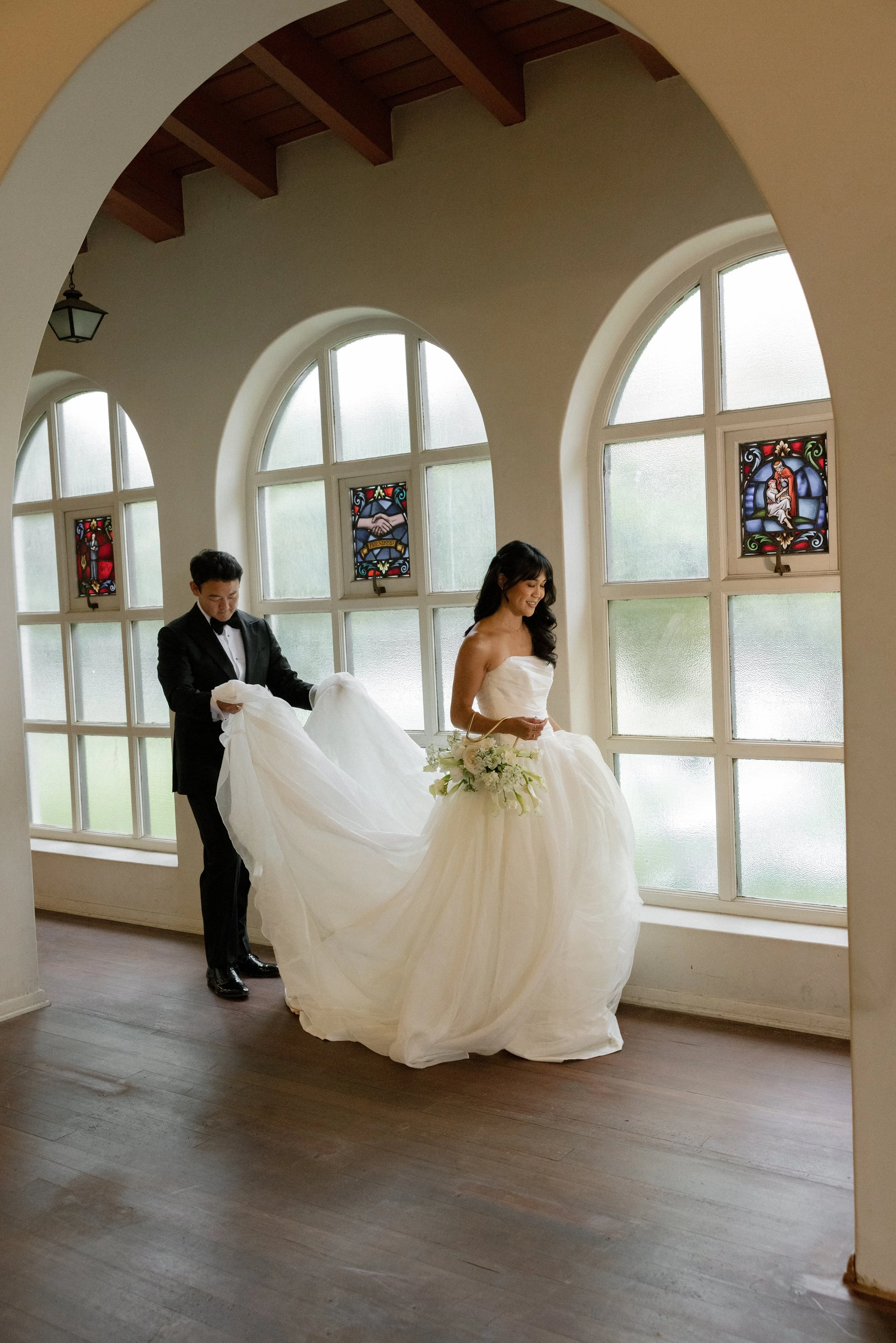 A bride in a strapless wedding dress holding a bouquet of white flowers, and a groom in a tuxedo, adjusting the bride's dress in front of large arched windows with stained glass details.