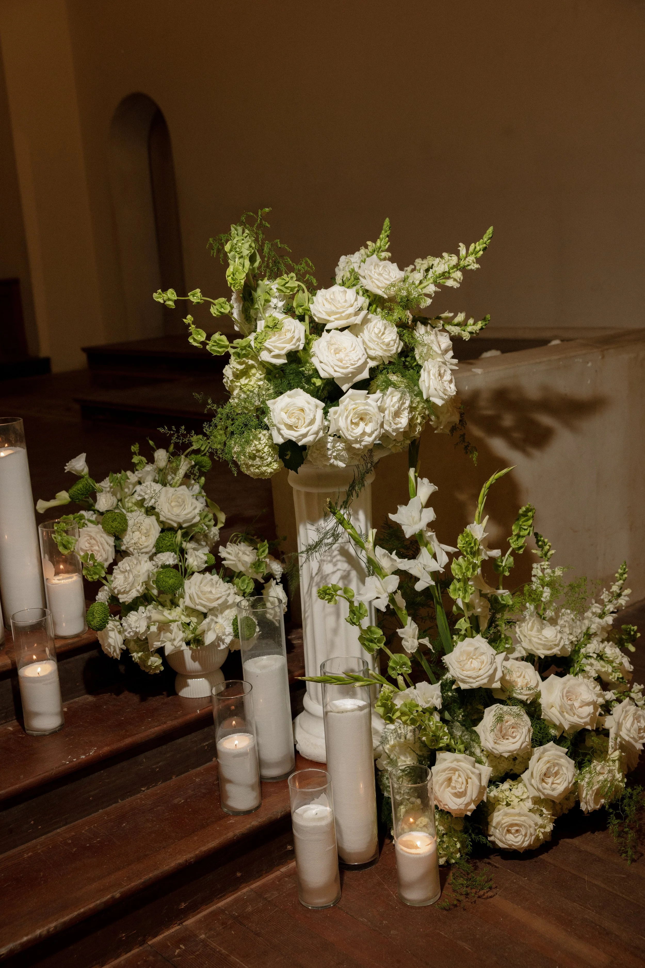 White flower arrangement on a table with candles, set on wooden stairs against a plain wall.