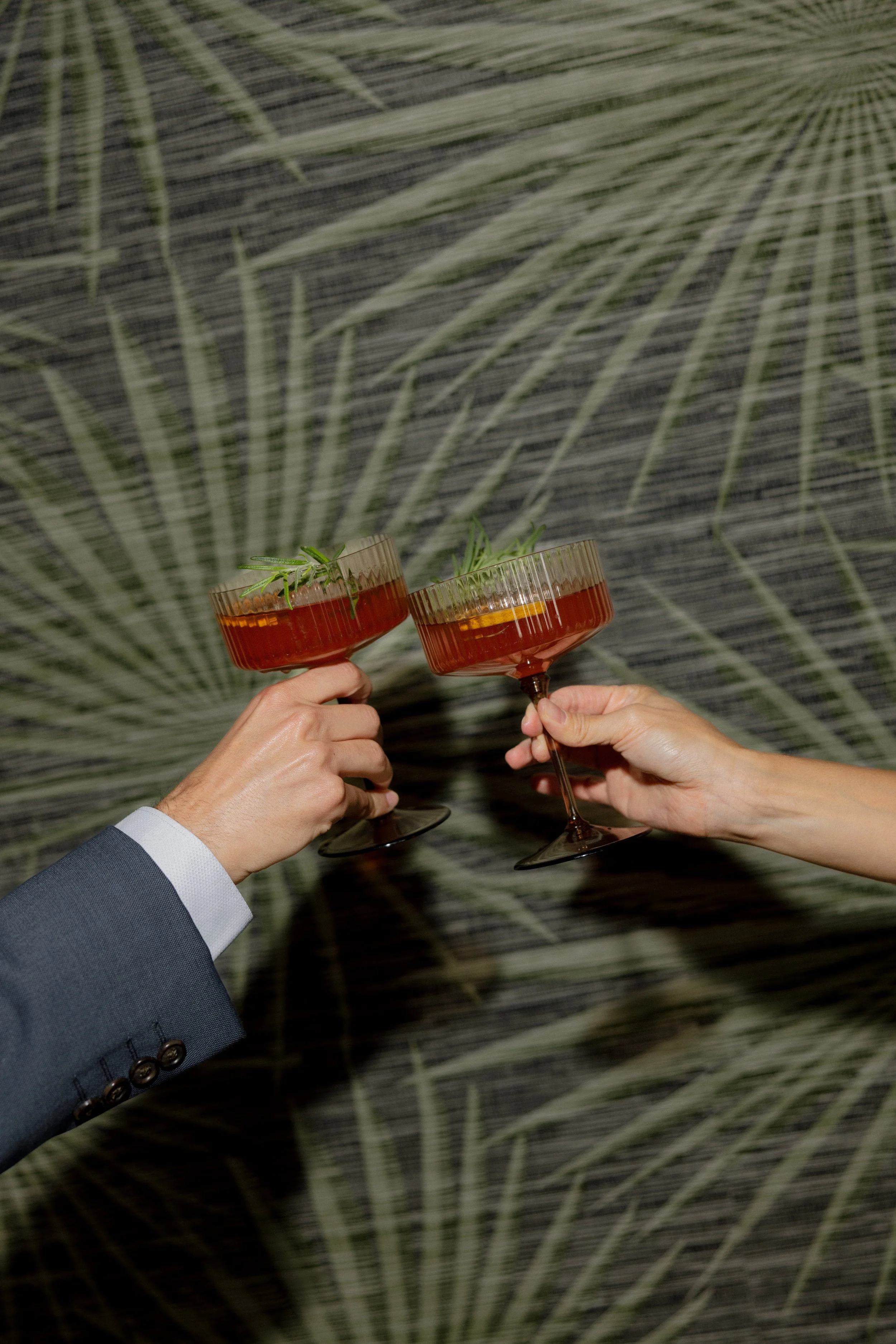 Two people holding cocktail glasses with orange drinks, garnished with herbs, against a background with a large palm leaf pattern.