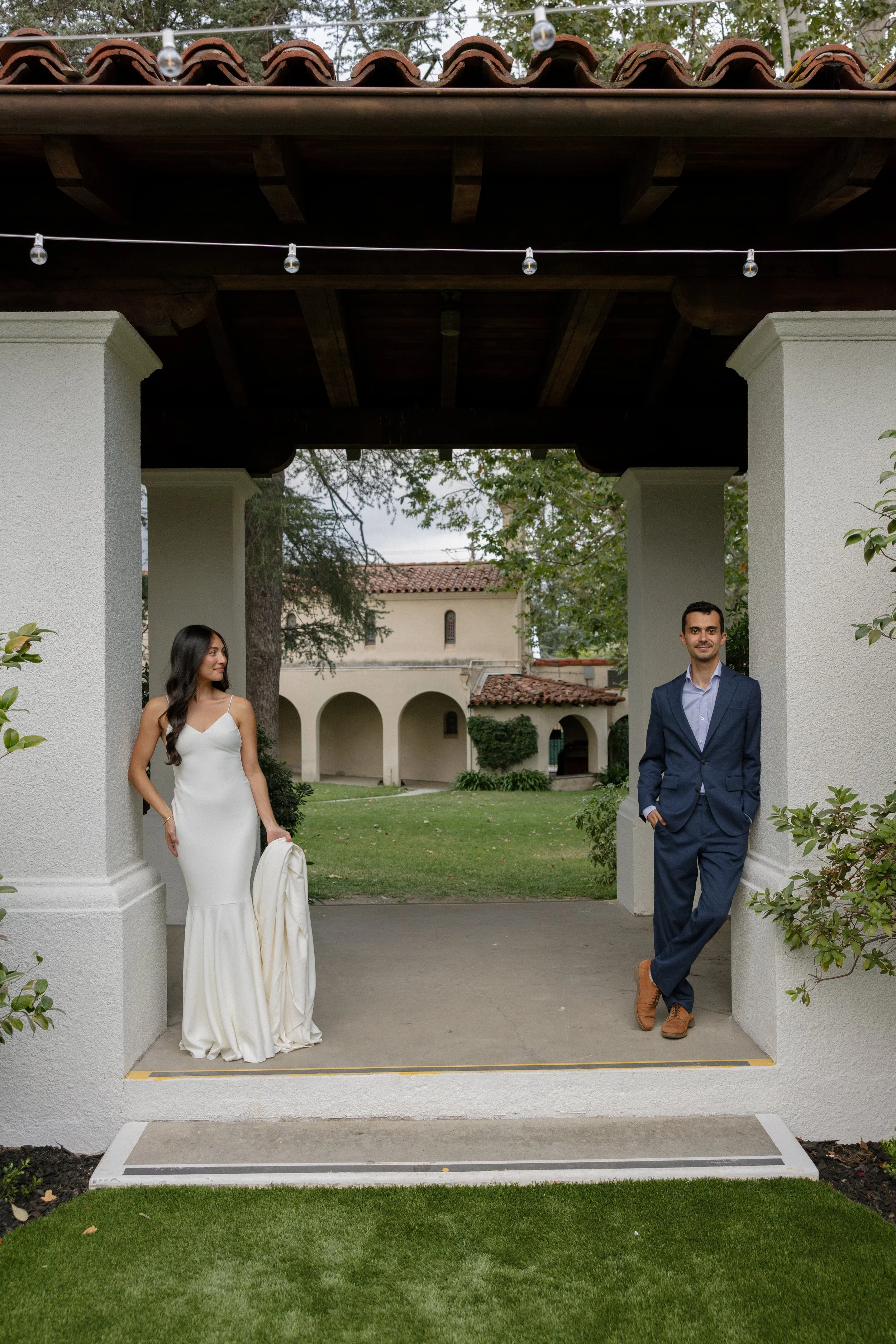 A woman in a white wedding dress and a man in a blue suit are standing under a white archway in a garden, with a building and trees in the background.