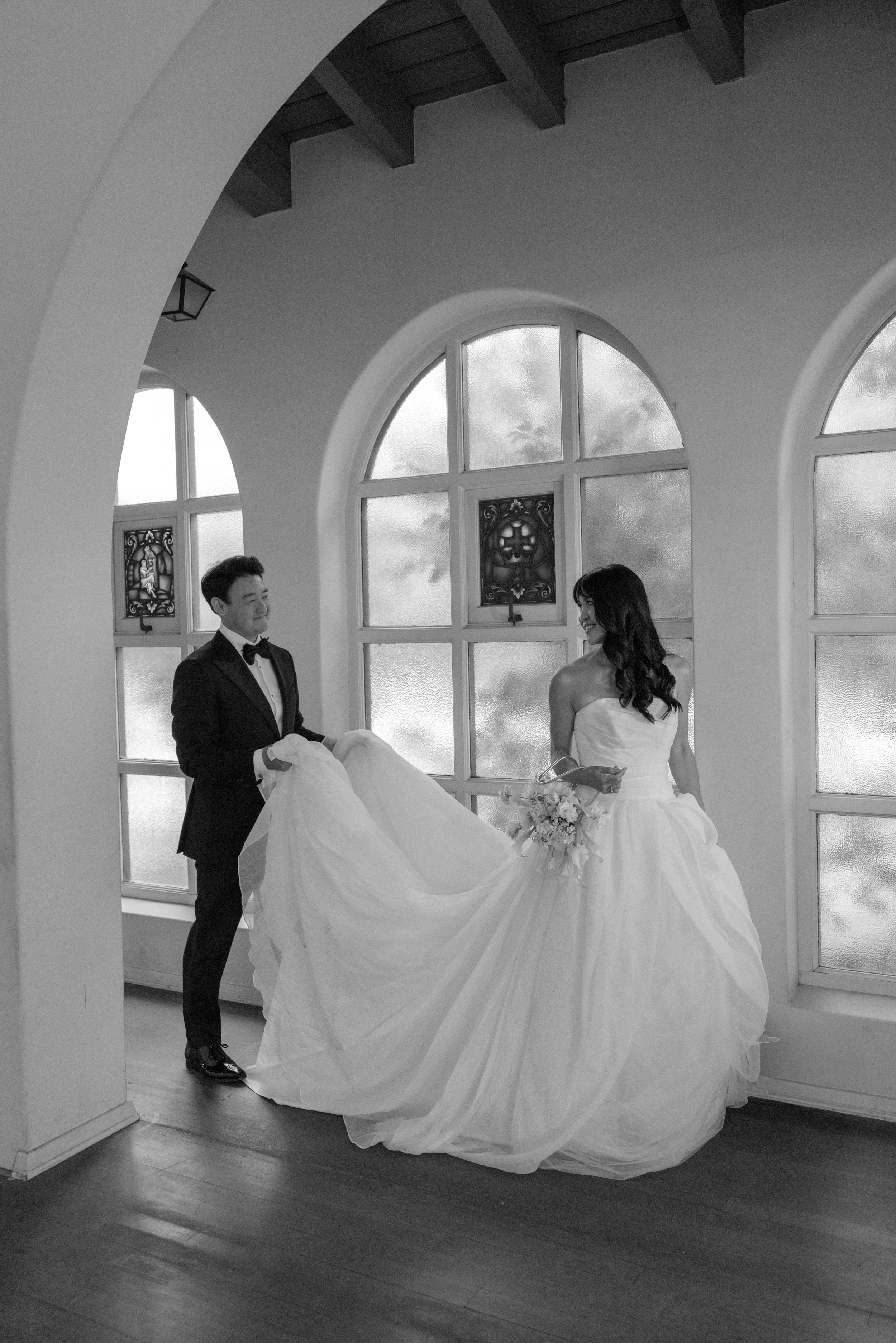 A bride and groom stand inside a room with large arched windows and wooden beams. The groom, dressed in a tuxedo, holds the bride's gown, while the bride, in a strapless wedding dress, holds a bouquet of flowers and smiles at the groom.