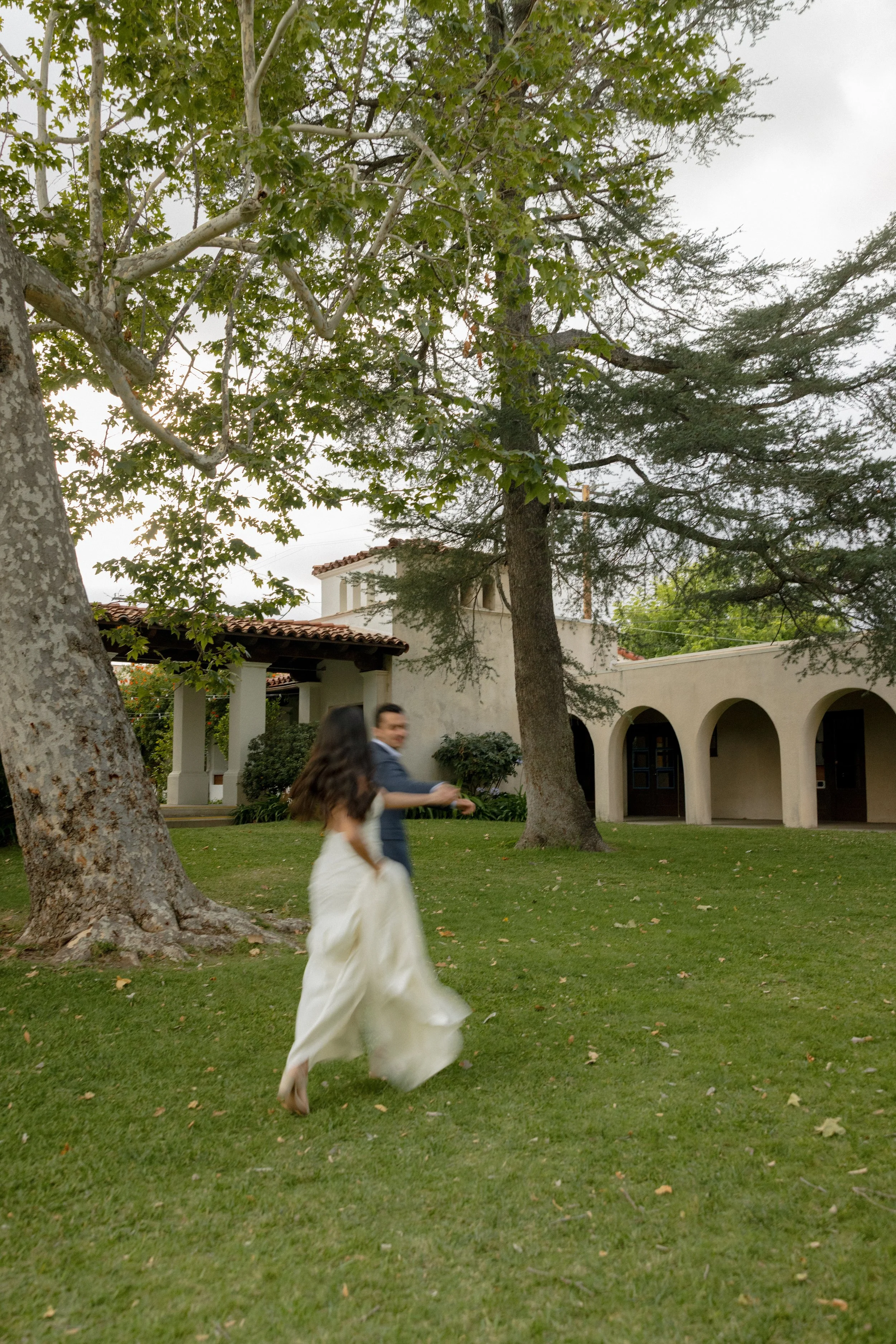 A woman in a white dress running across a grassy yard with large trees and a building with arches in the background.