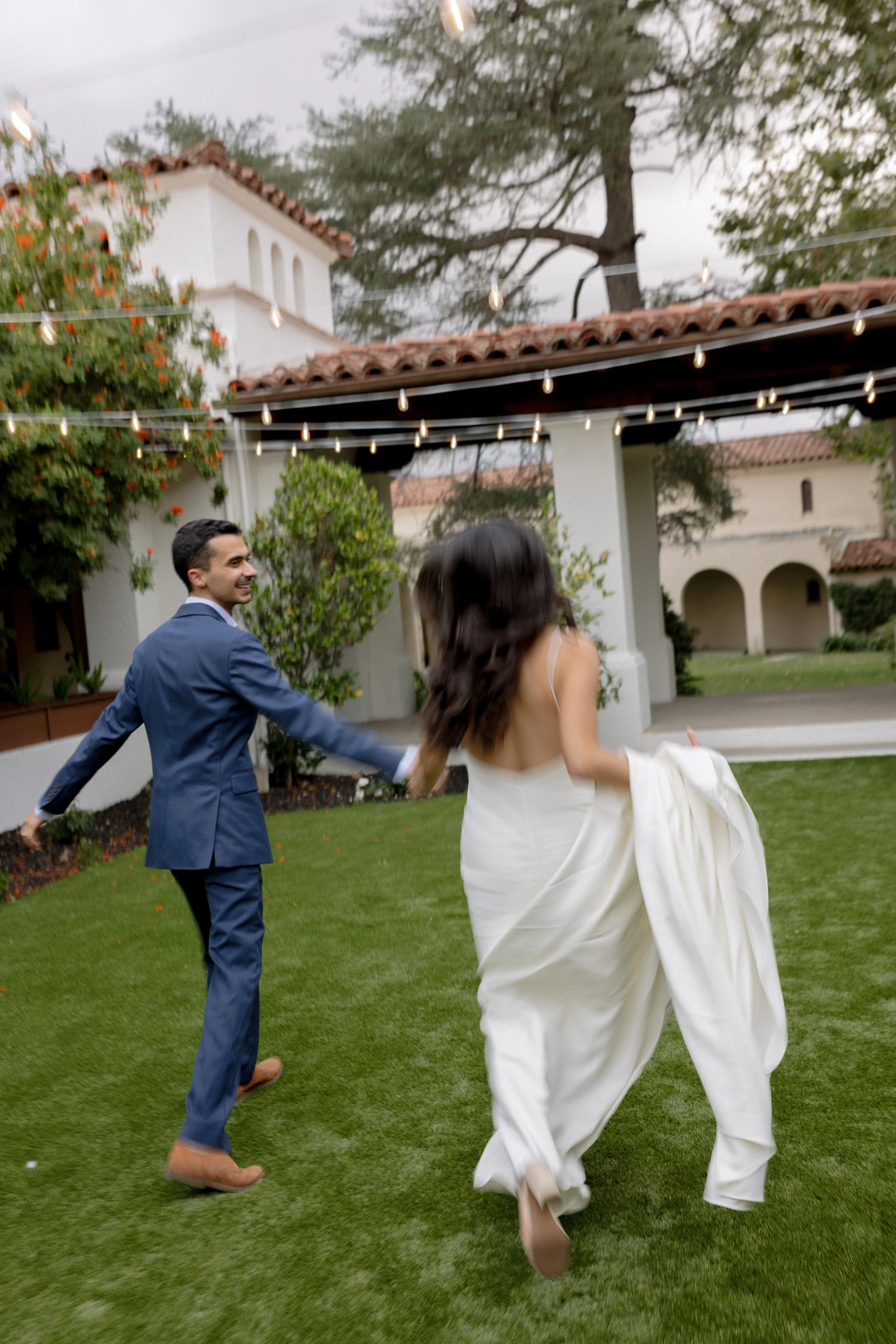 A man in a blue suit and a woman in a white dress are dancing and holding hands in a backyard with string lights overhead and greenery around.