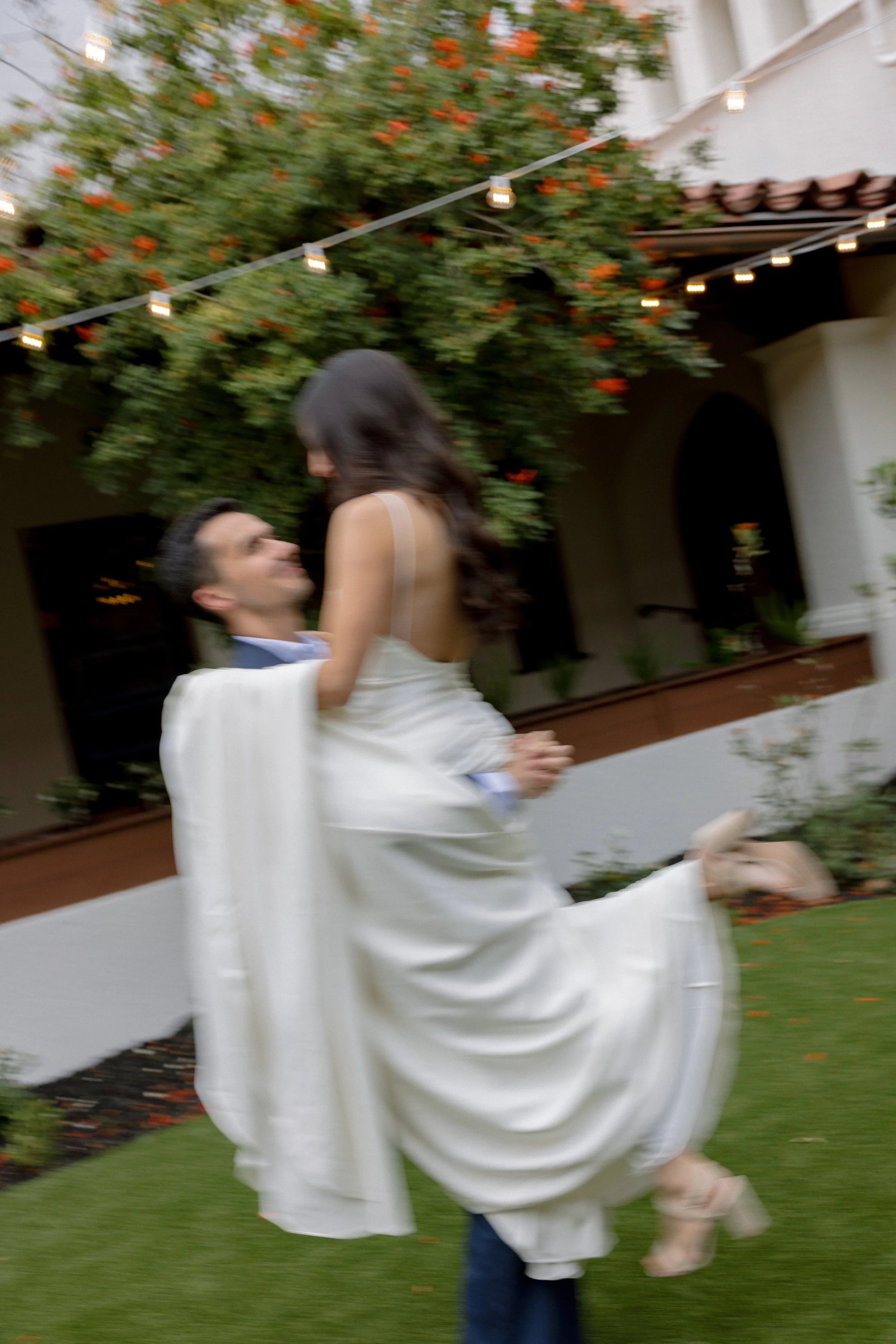 A blurred photo of a couple dancing outdoors with string lights and a flowering tree in the background. The man is lifting the woman, and she is wearing a white dress.