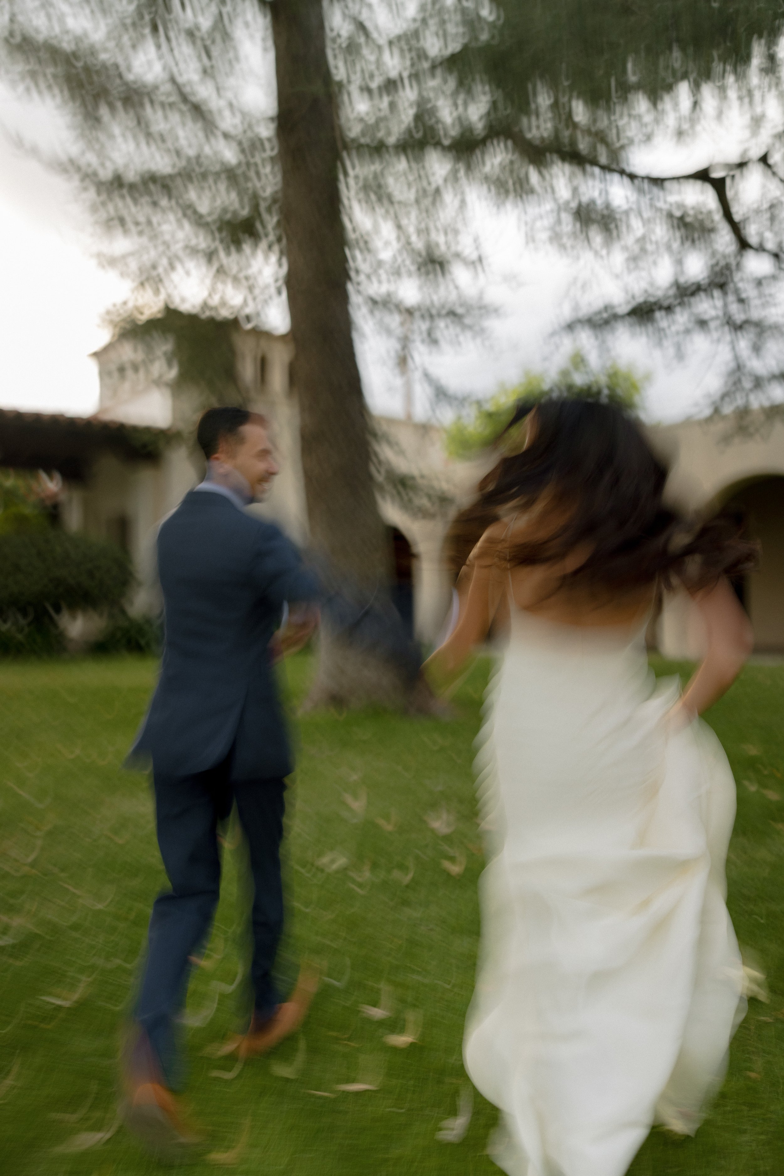 A blurred image of a bride and groom holding hands outdoors, with a large tree and building in the background, and leaves swirling in the air.