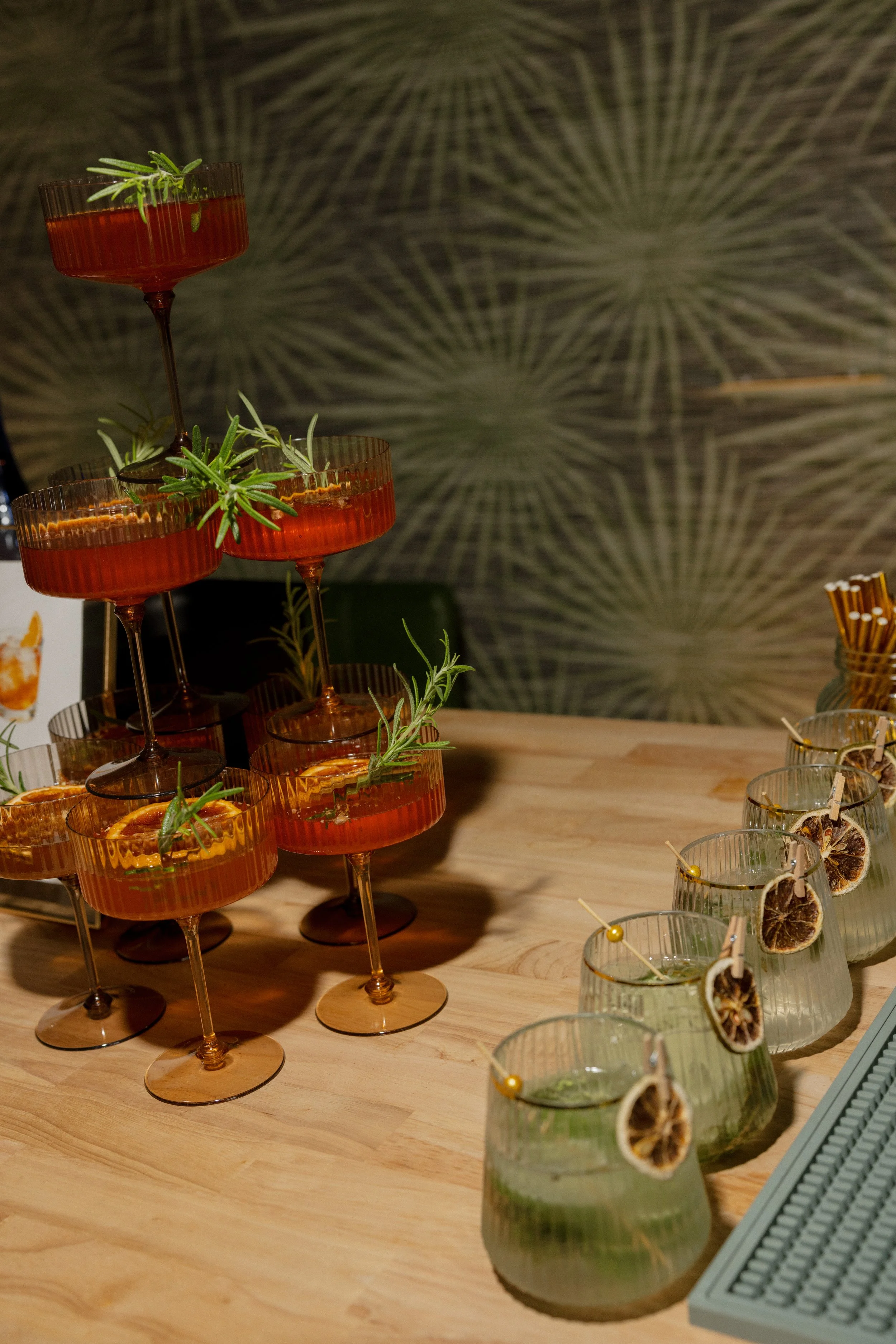 A display of cocktails in orange-colored glasses with sprigs of rosemary and slices of dried citrus on a wooden surface, with additional clear drinks garnished with dried citrus slices in the foreground.