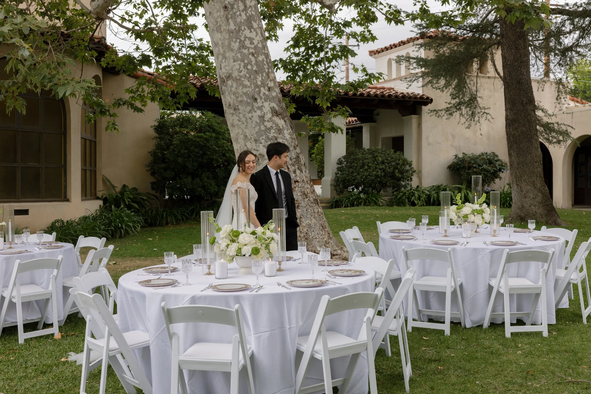 Outdoor wedding setup with round tables covered in white tablecloths, white chairs, floral centerpieces, and a bride and groom standing near a large tree in front of a beige stucco building.
